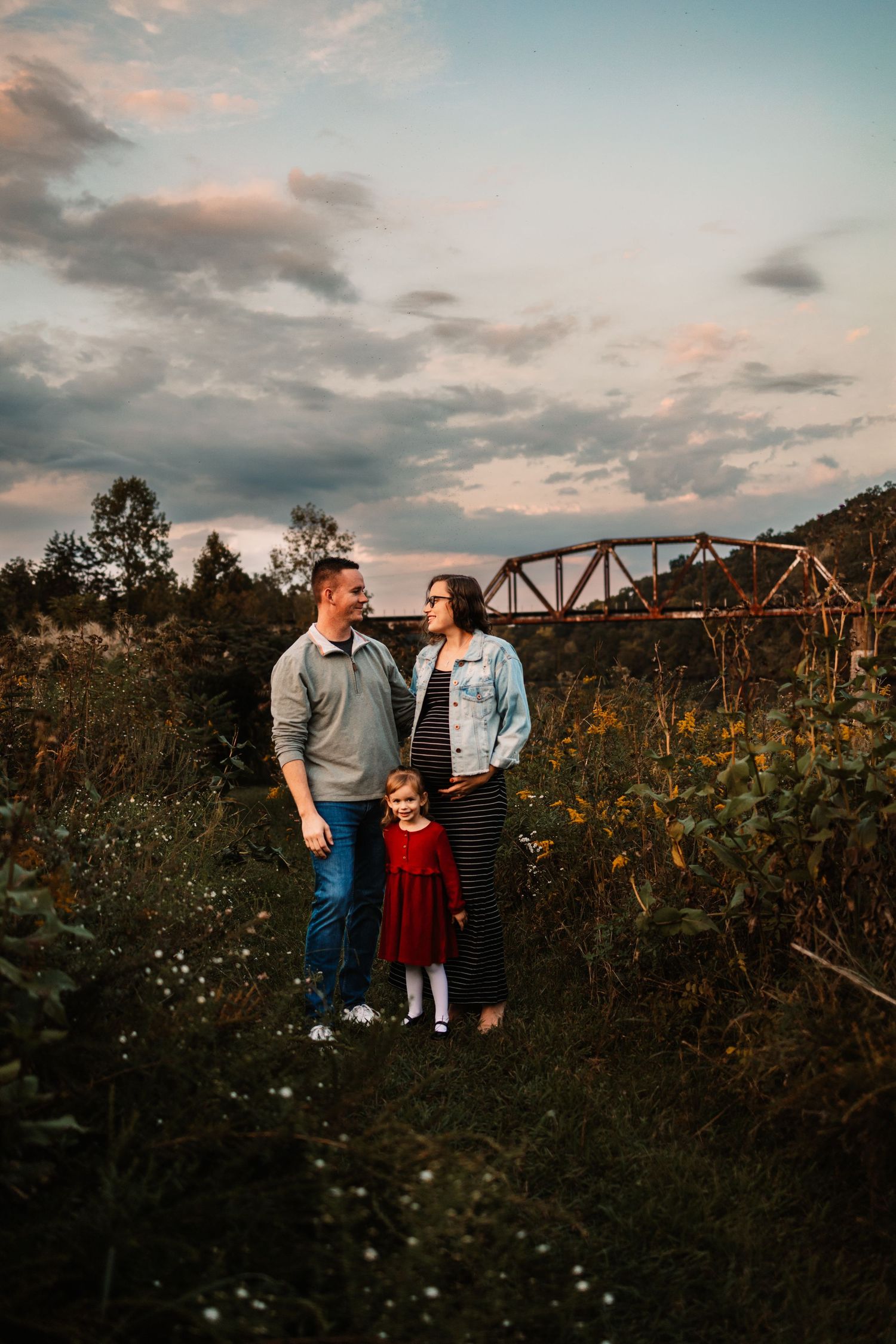 A family of three poses in a field at sunset, with a rustic bridge backdrop, mother in denim jacket showing a baby bump.
