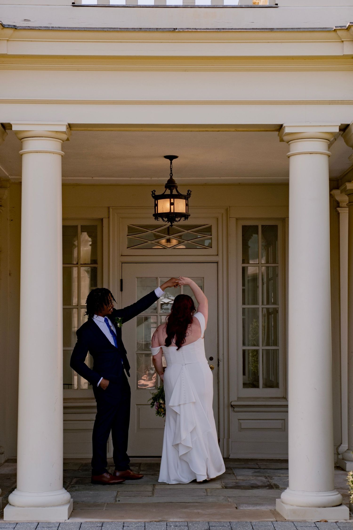 Wedding party celebrates with dancing and laughter under elegant white columns on a covered porch.