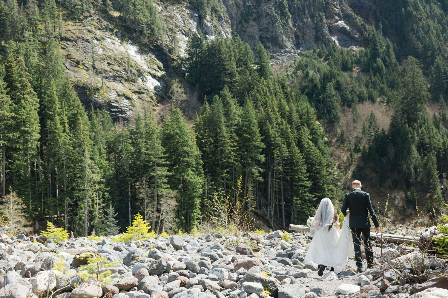 Bride and groom walking on rocky terrain surrounded by tall evergreen trees and mountains on their wedding day.