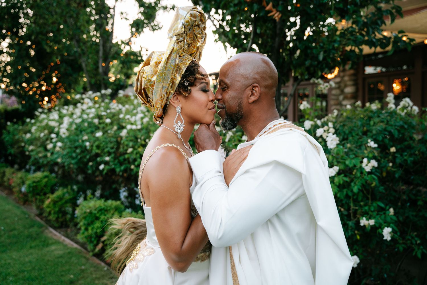 Couple in traditional white and gold attire walking through lush green garden during outdoor wedding ceremony.