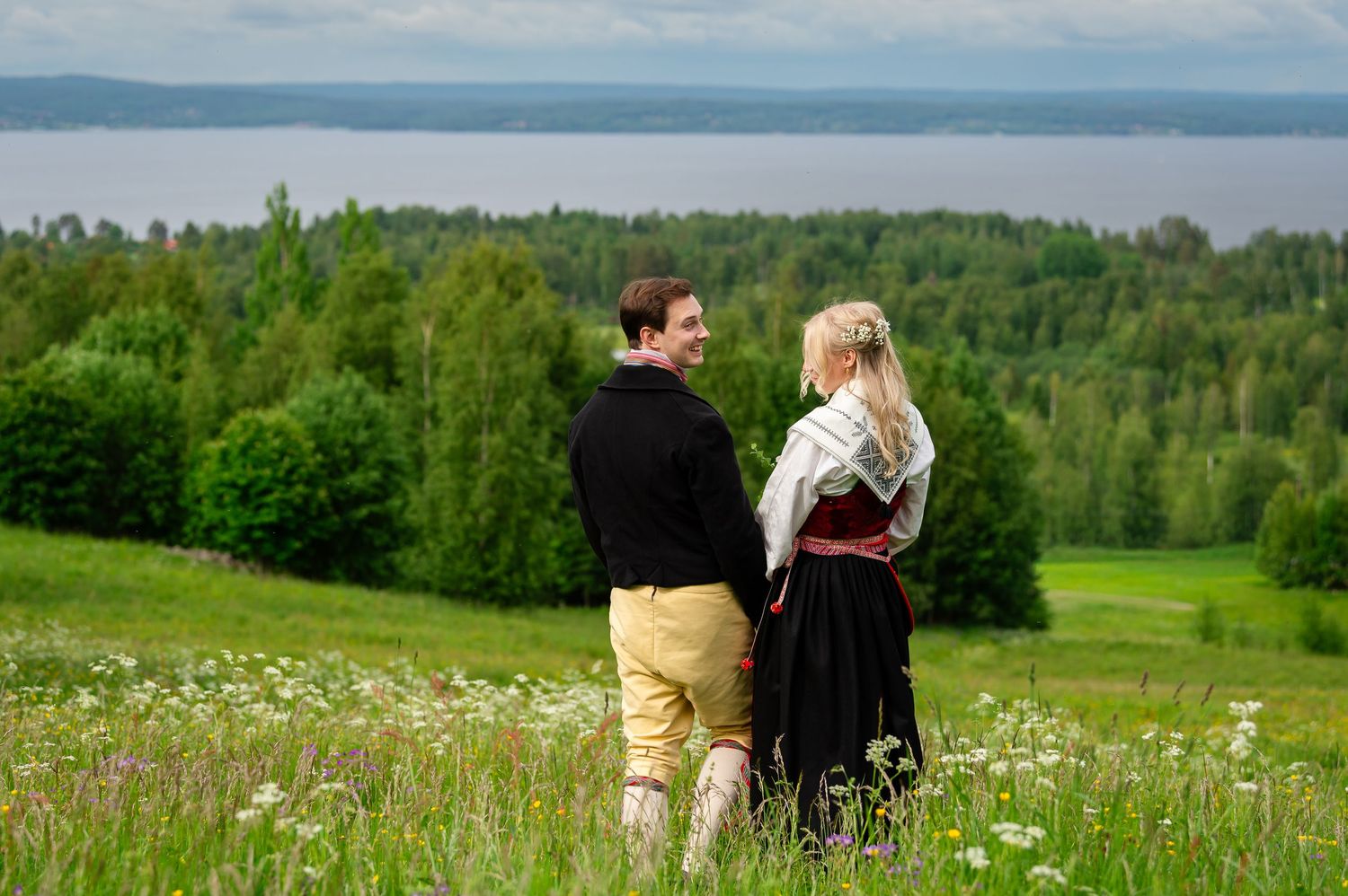 Couple in traditional attire holding hands in a lush green meadow with forest and lake views.
