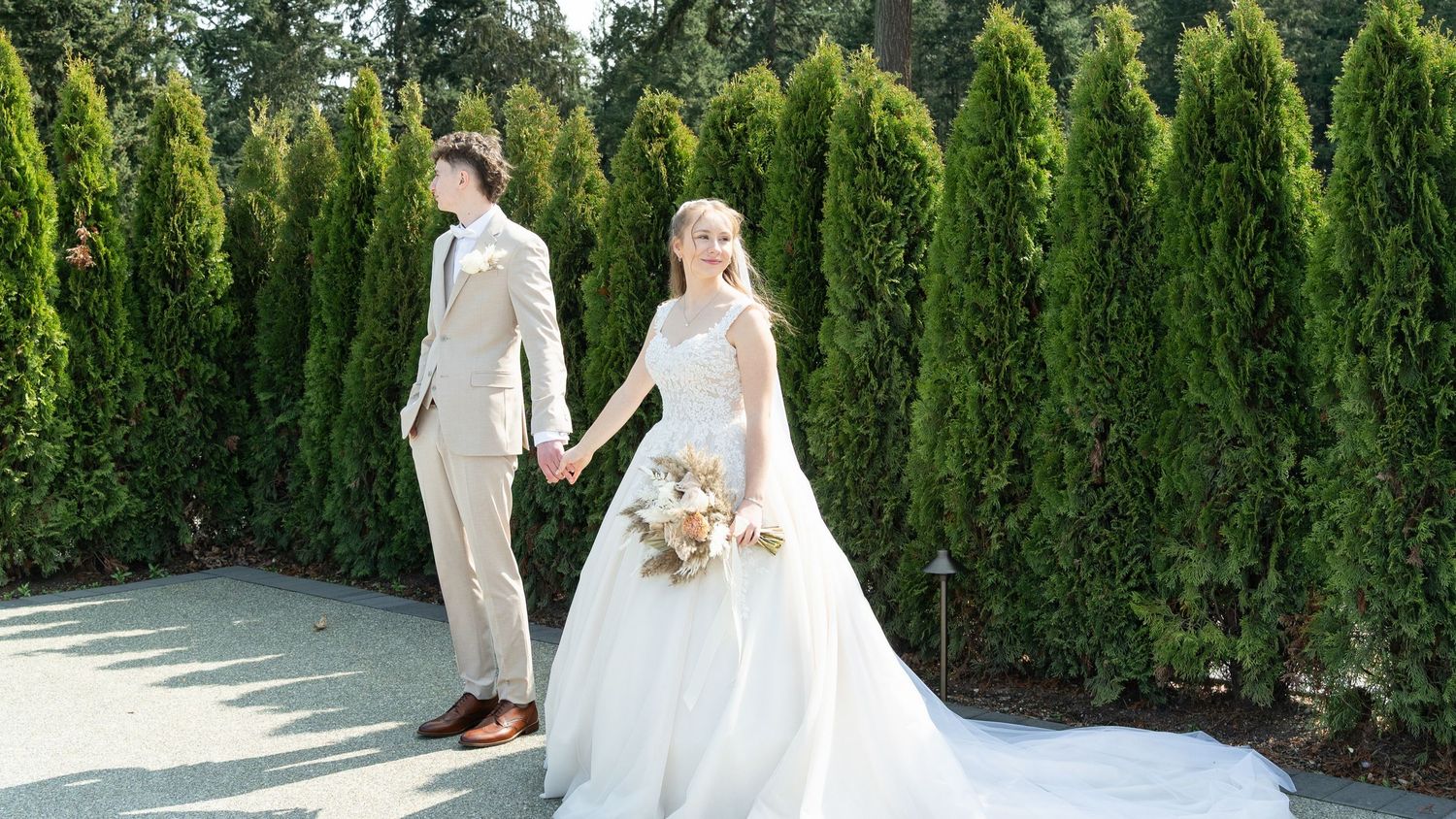 A bride in a flowing white gown and groom in a beige suit hold hands outdoors, surrounded by tall evergreen trees.