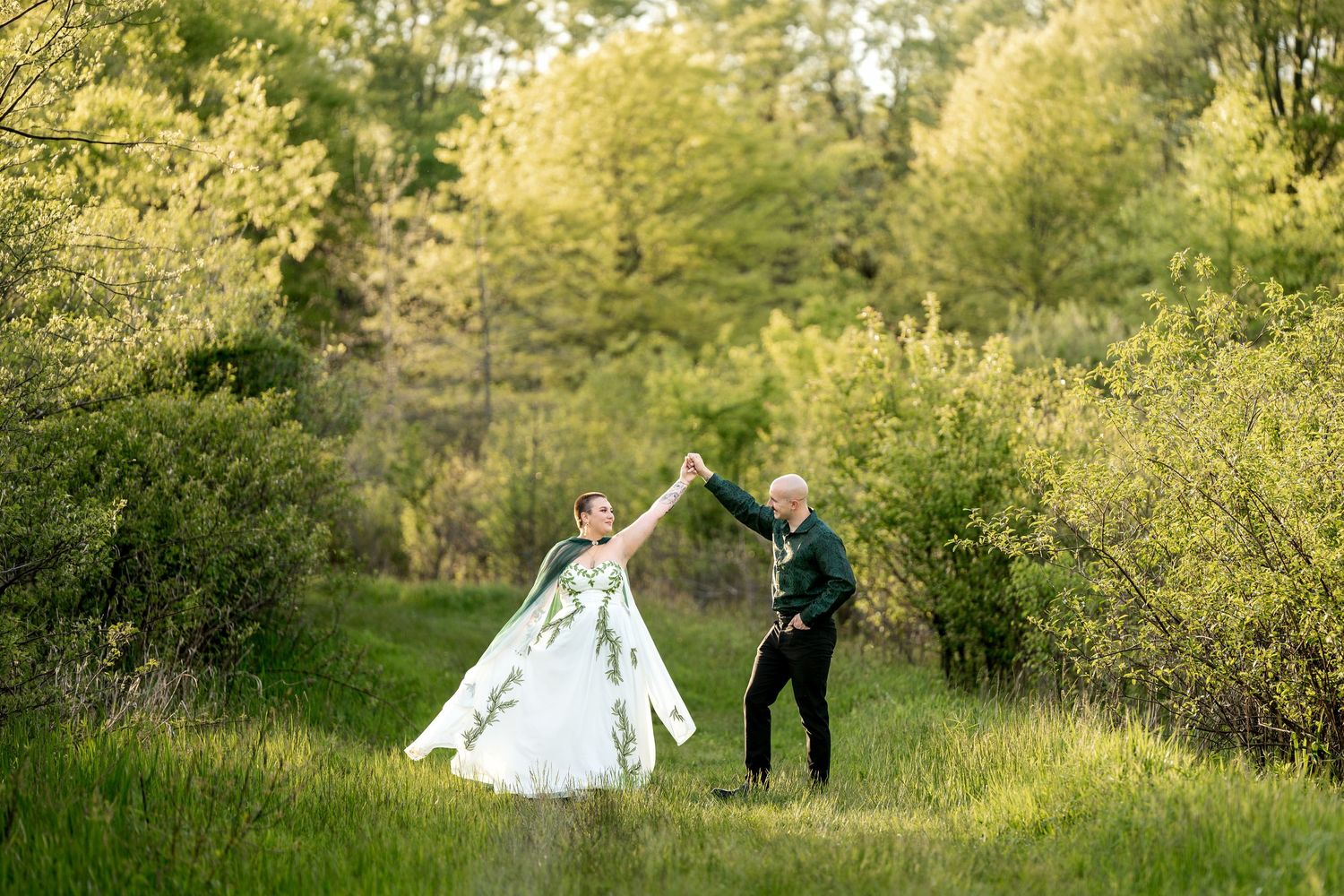 Bride and groom dancing in a lush green meadow surrounded by trees, bride wearing a floral white gown.