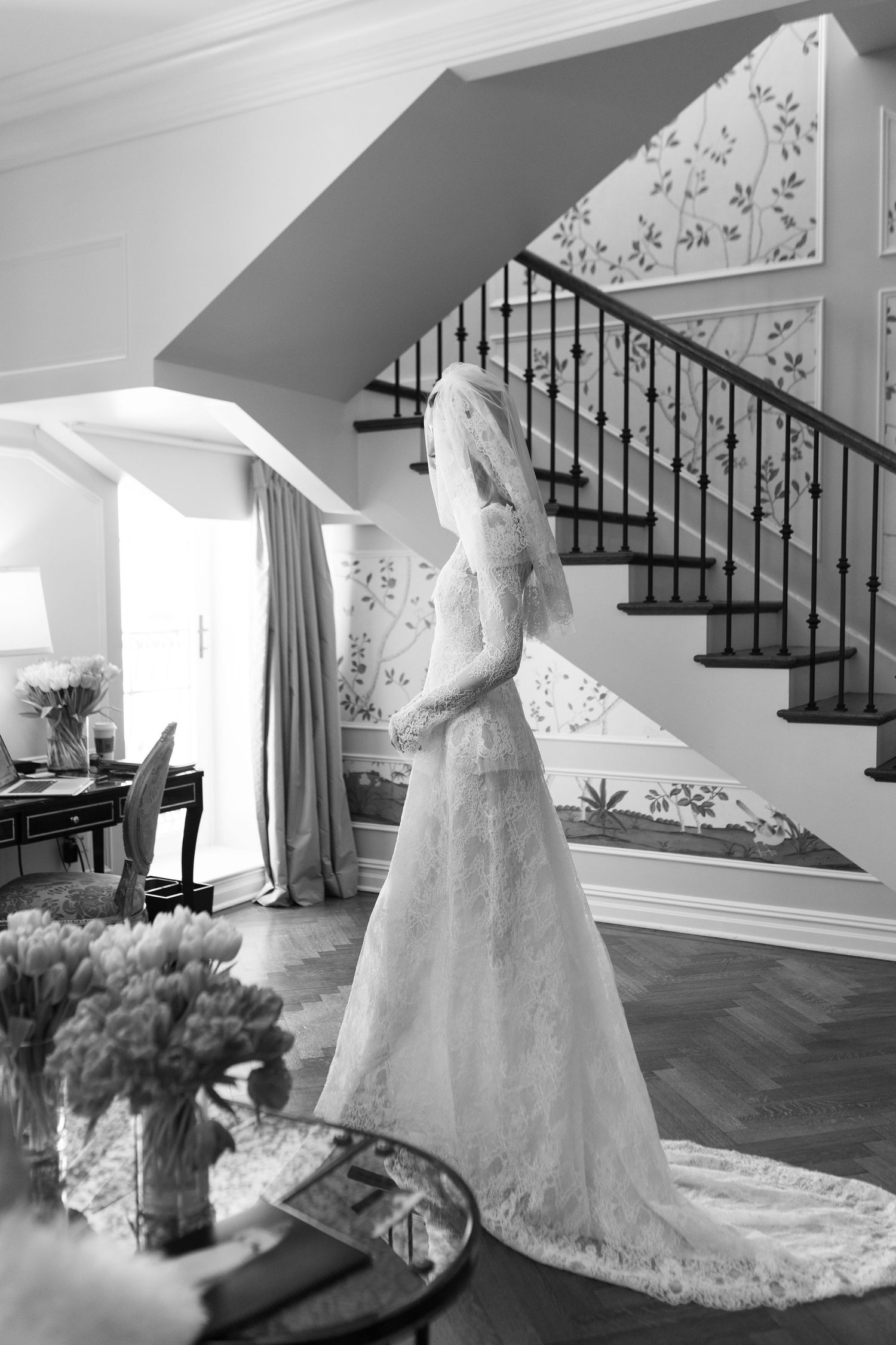 Blonde woman in a white lace bridal gown poses against a floral wallpaper backdrop in an elegant bridal boutique.