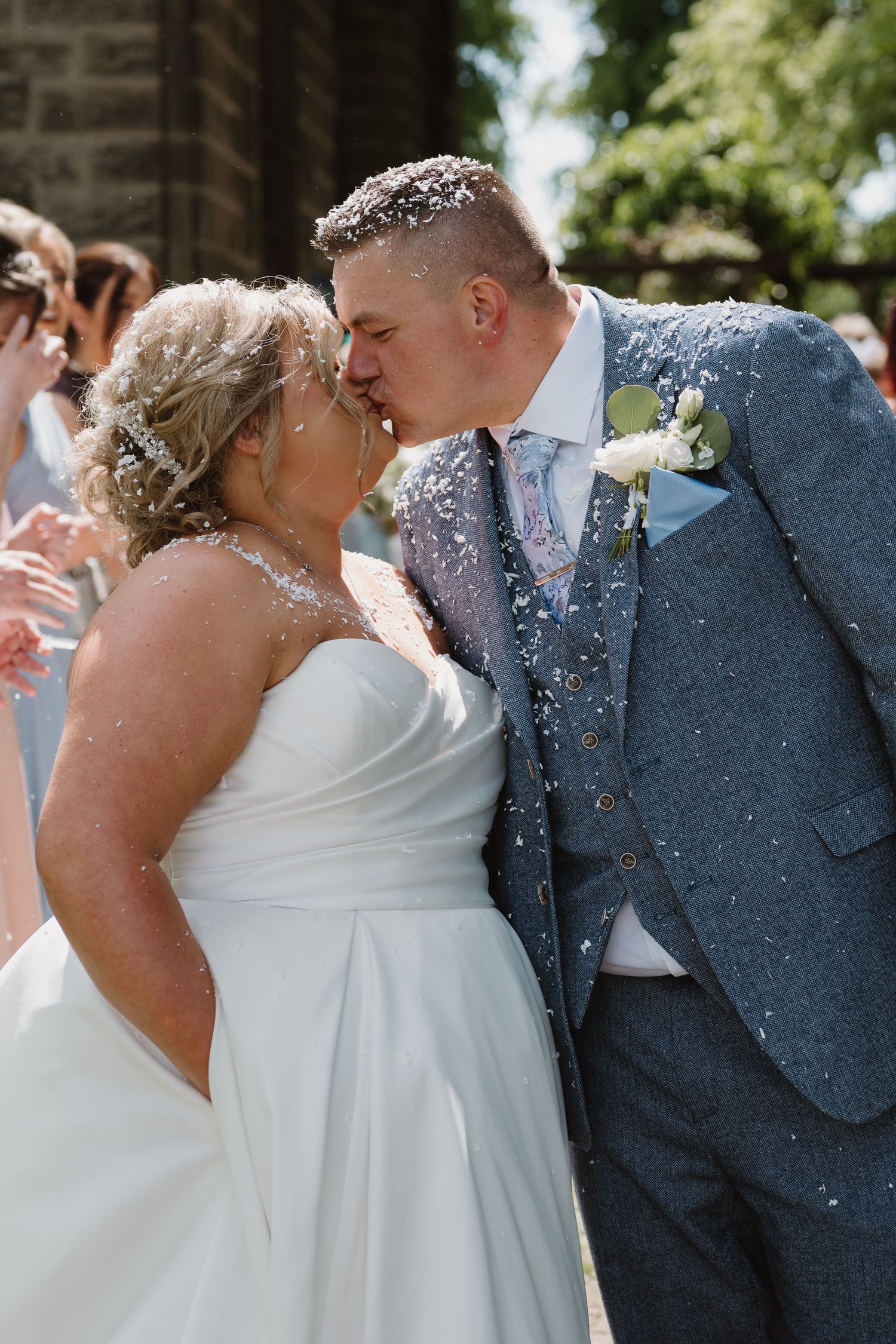 Bride and groom share a kiss outdoors as confetti falls around them, bride in white strapless gown, groom in blue tweed suit.