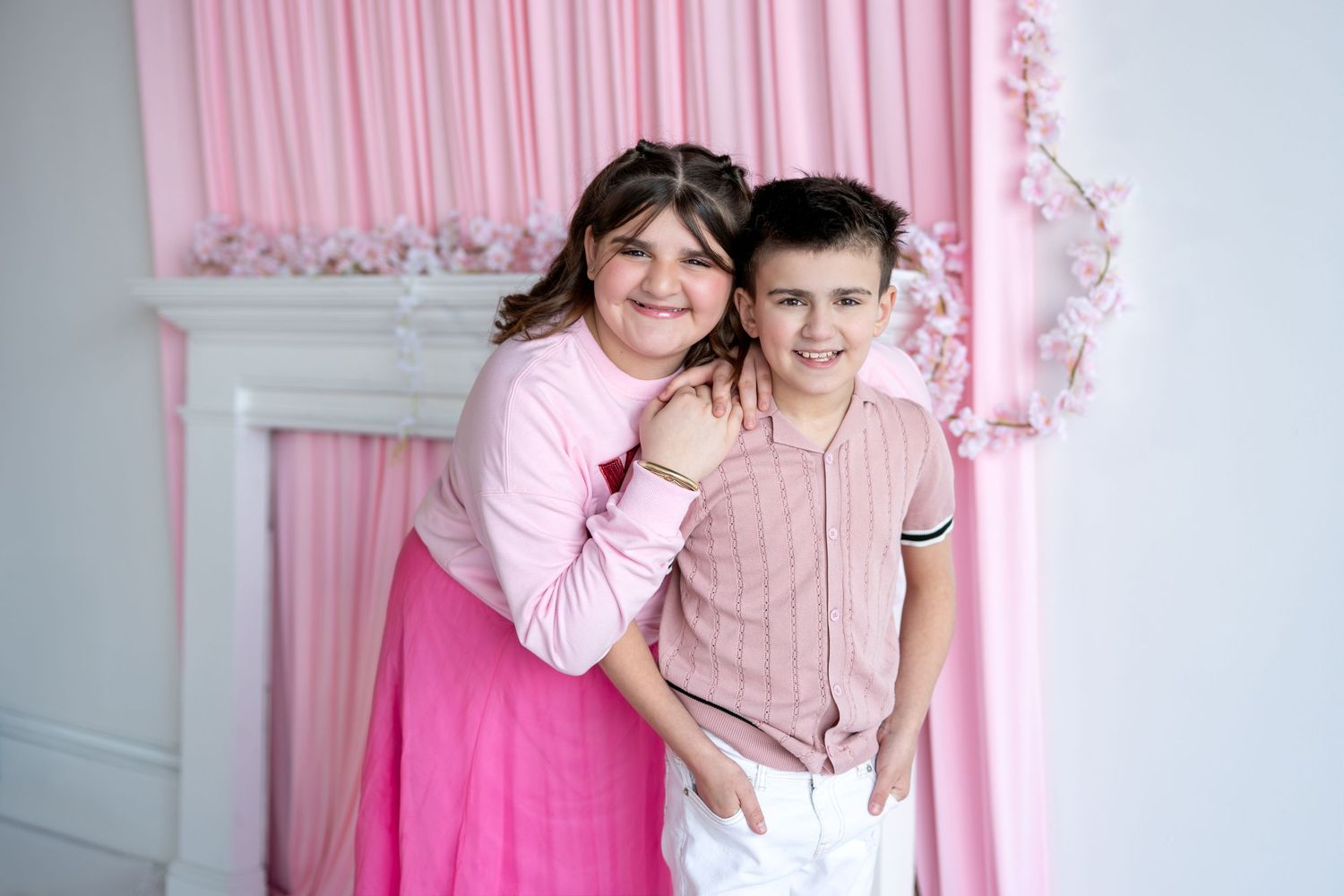 Two children smiling together, girl in pink outfit and boy in pink shirt posing before pink floral backdrop.