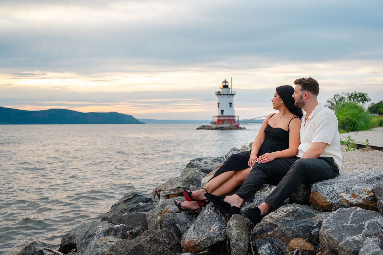 Man proposing on one knee to woman in black dress by lighthouse on waterfront at sunset.