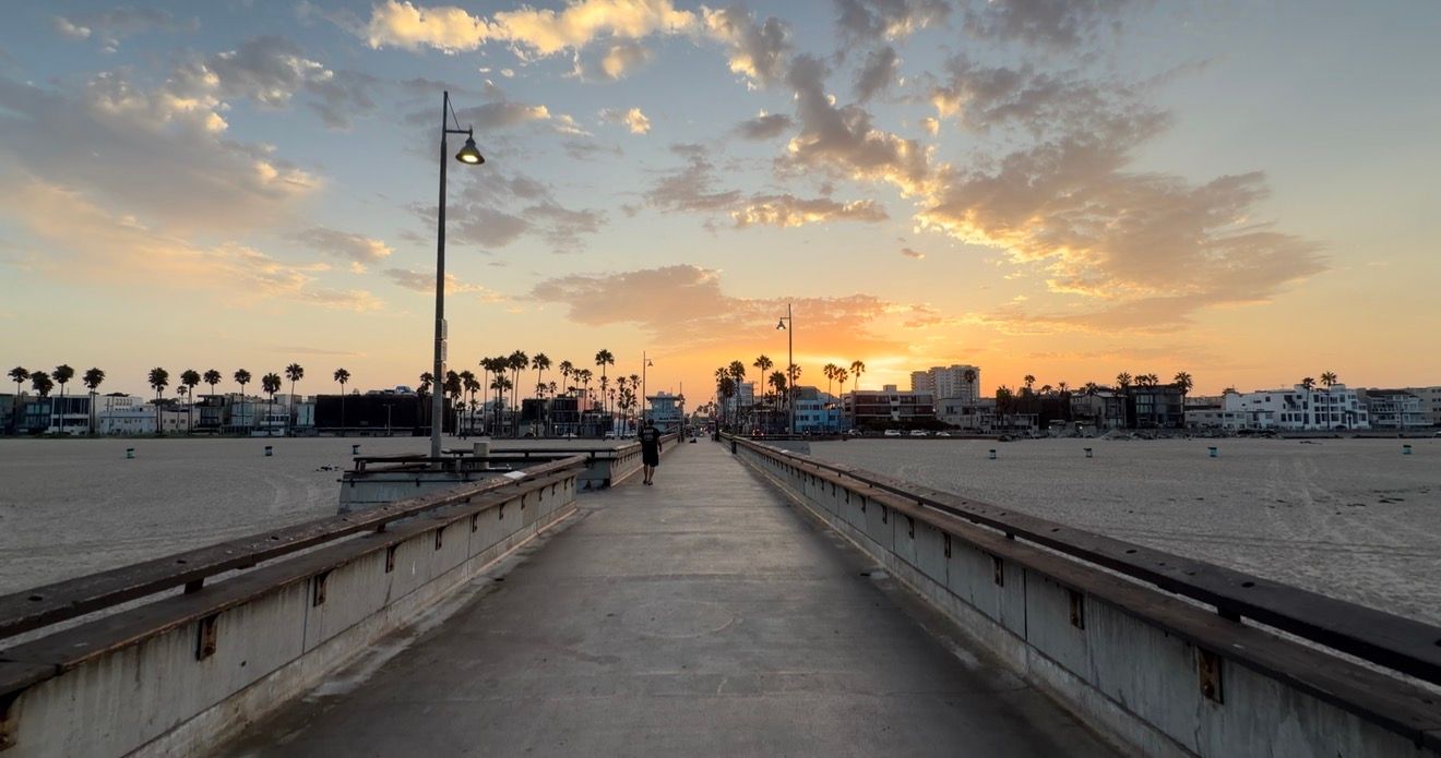 Scenic beachside pier at sunset with palm trees, golden sky, and people walking toward the horizon.