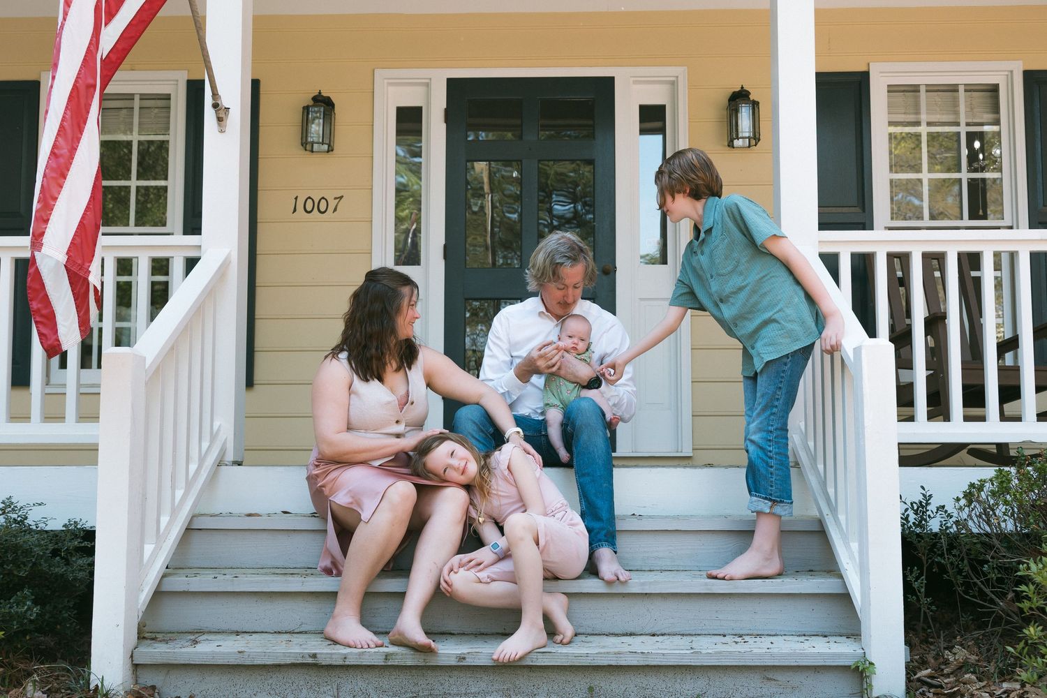 Family of five relaxing on front porch steps of yellow home with American flag, sharing a warm moment together.