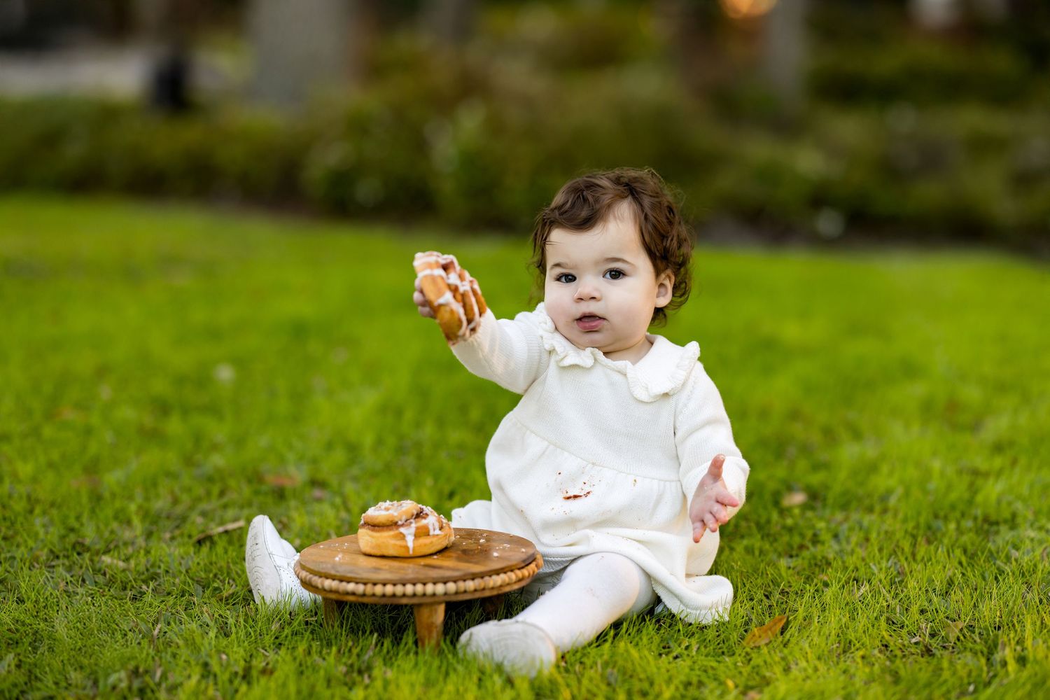 Small child in white outfit sits on grass with basket of bagels during outdoor photo session.