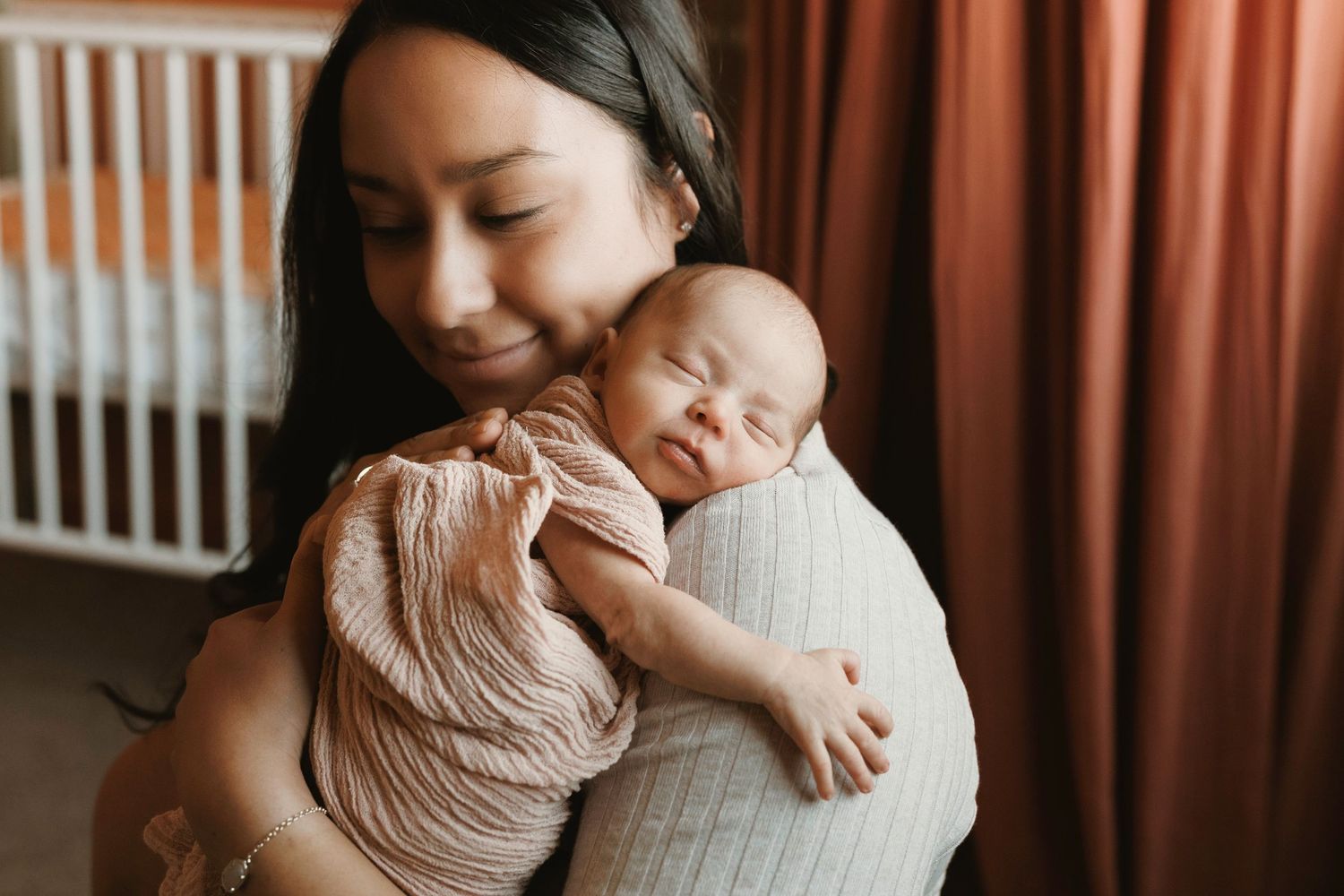Mother tenderly holding sleeping swaddled newborn baby against her shoulder.