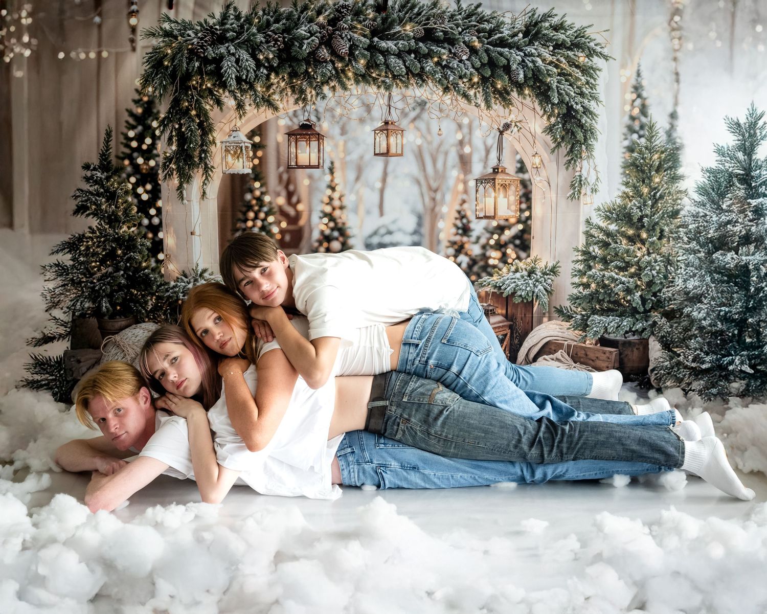 A cozy winter scene with people lying on white fur blankets surrounded by evergreen trees and twinkling lights.