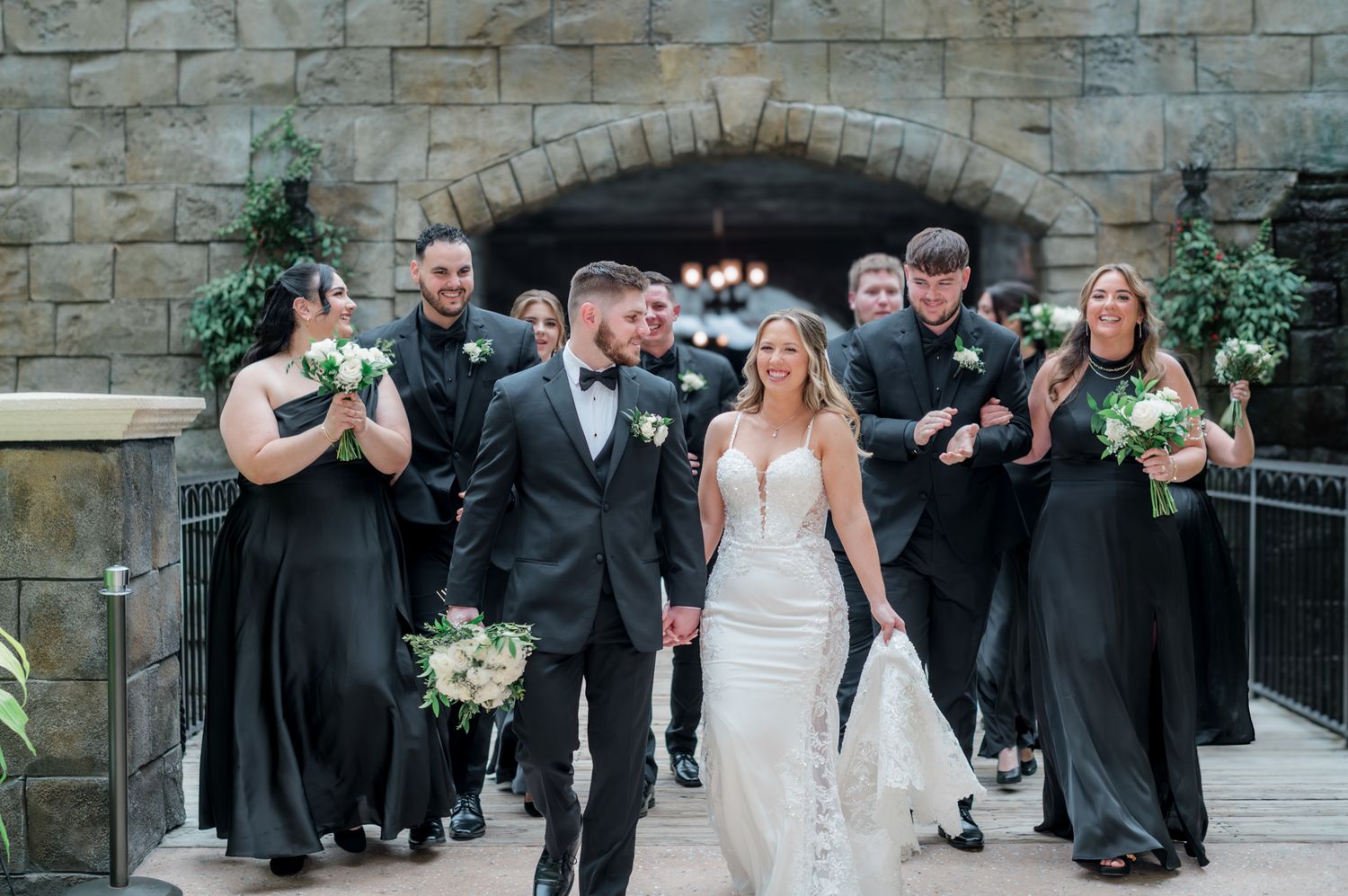 Bride and groom walk hand-in-hand with wedding party in black attire, holding white bouquets, under stone archway.