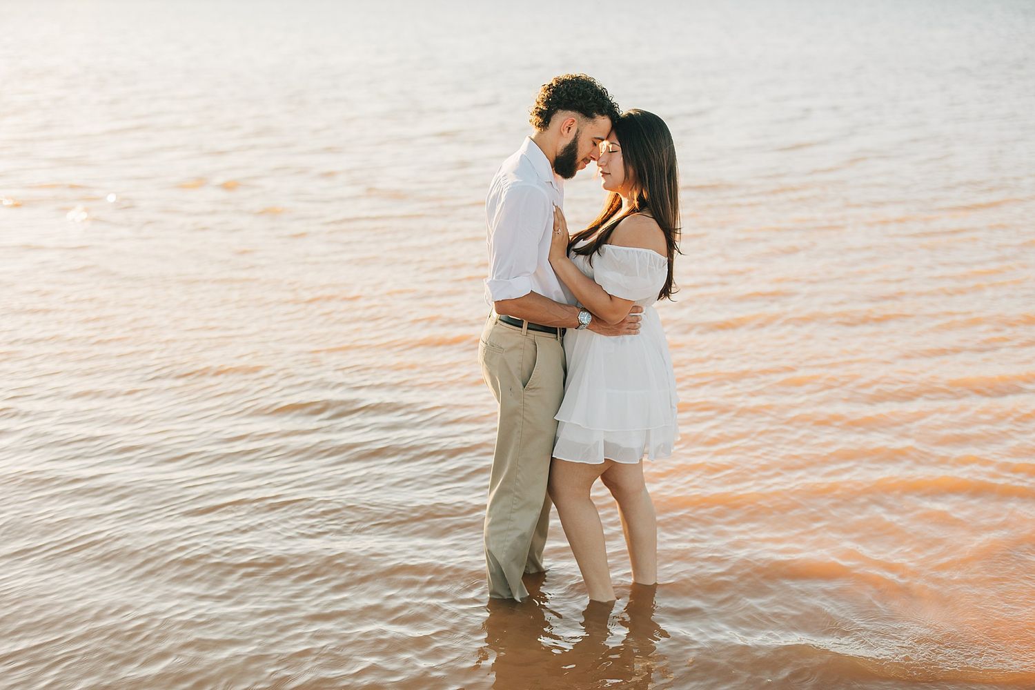 A romantic couple embraces while standing in shallow water at sunset on a beach.