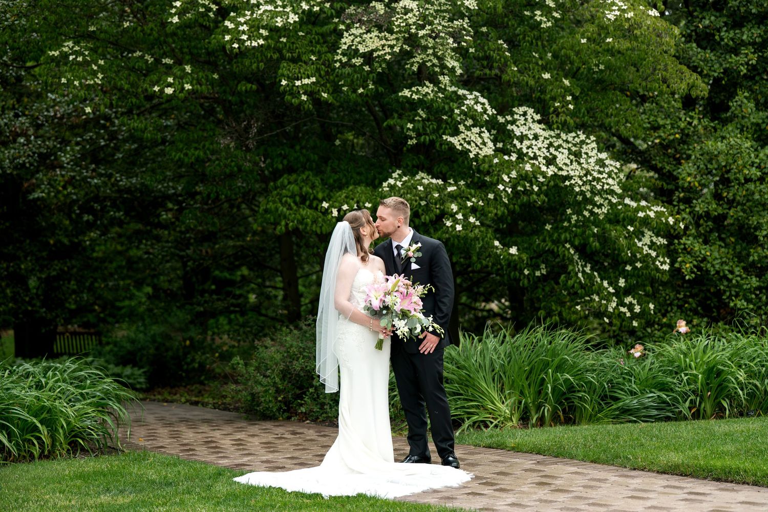 Bride and groom kissing in lush garden setting, bride in white gown with veil holding pink bouquet.