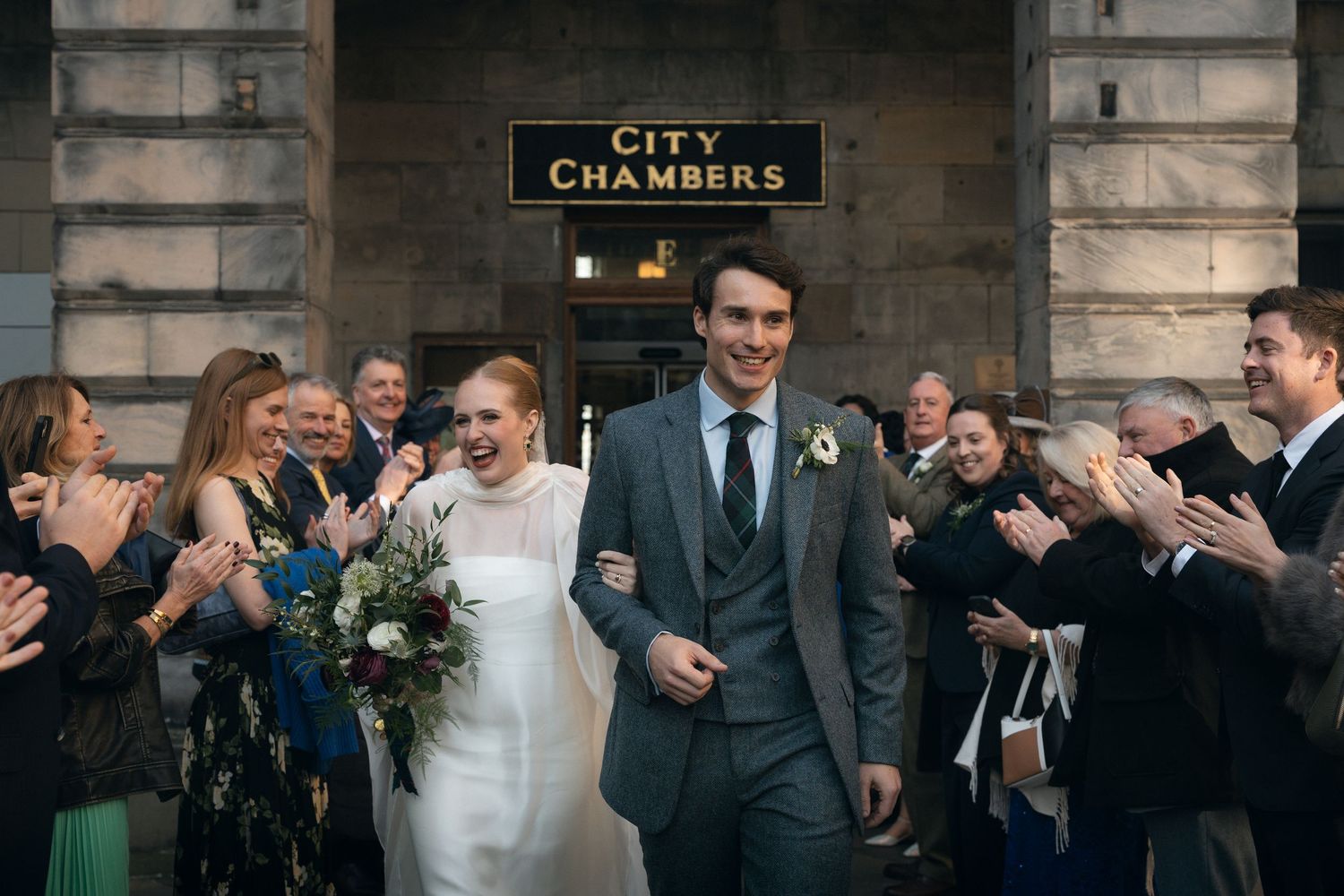 Wedding party celebrates with laughter outside historic City Chambers building in formal attire.