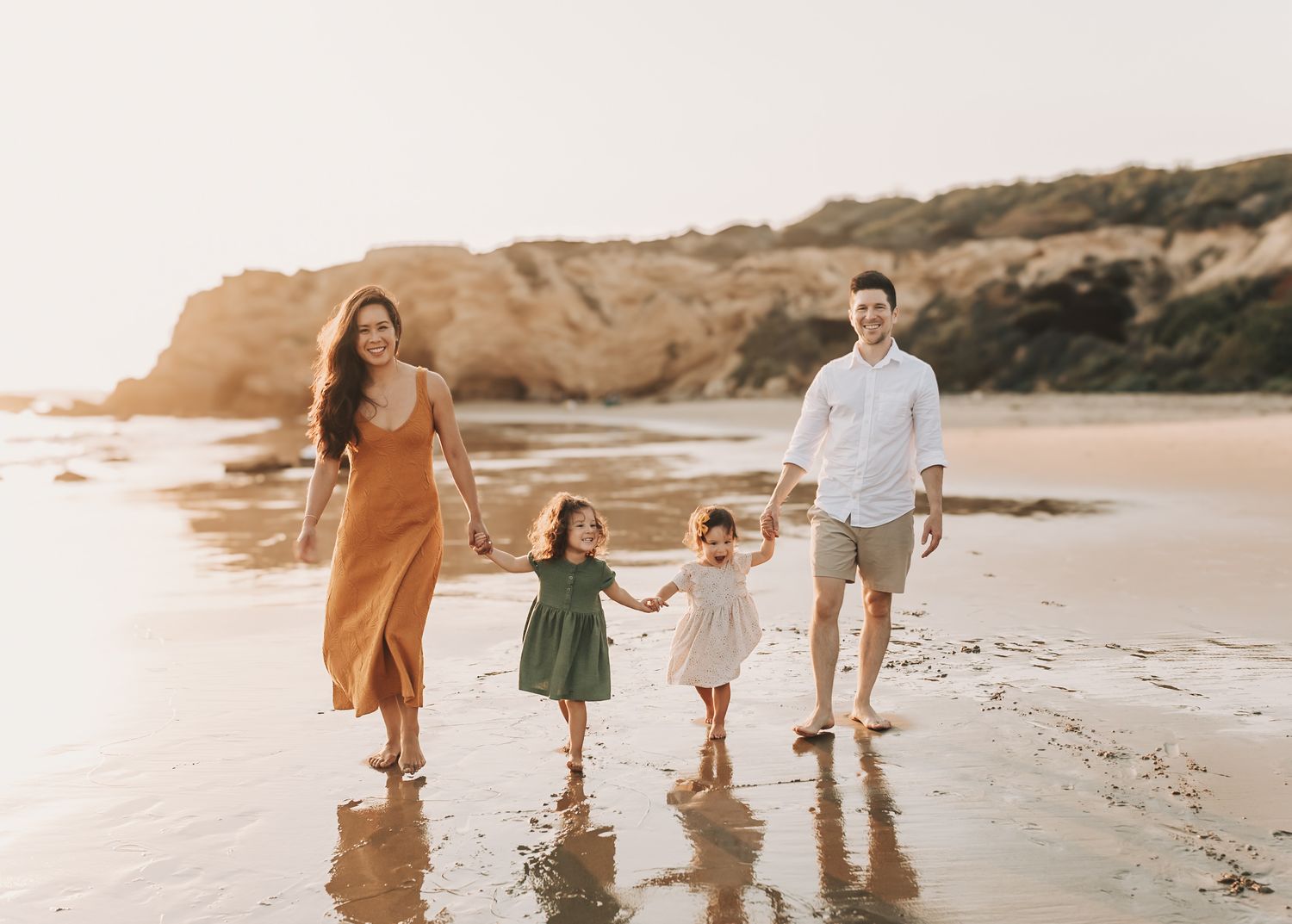 Family walking along sandy beach at sunset with cliffs in background and waves lapping at their feet.