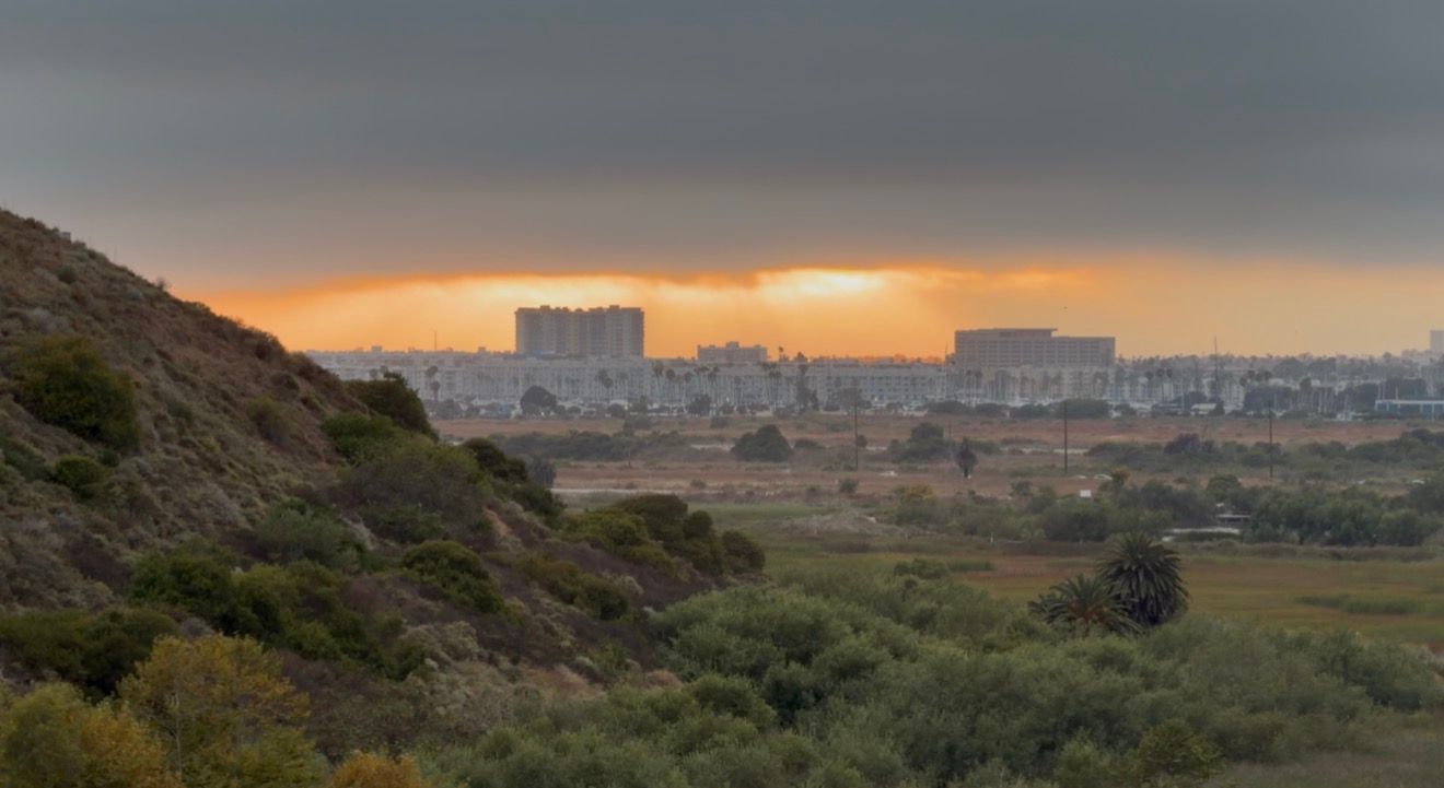 Panoramic sunset view over a city skyline with dramatic orange sky, green hills, and lush valley in the foreground.