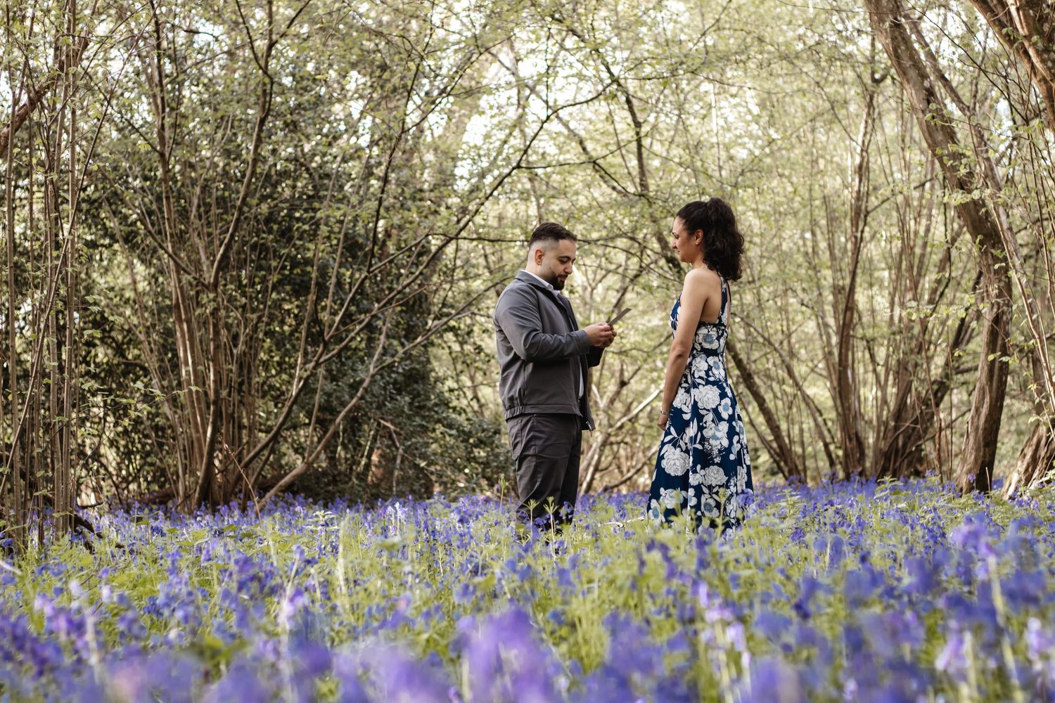 Couple standing together in a bluebell woodland field, surrounded by purple wildflowers and trees in soft natural light.