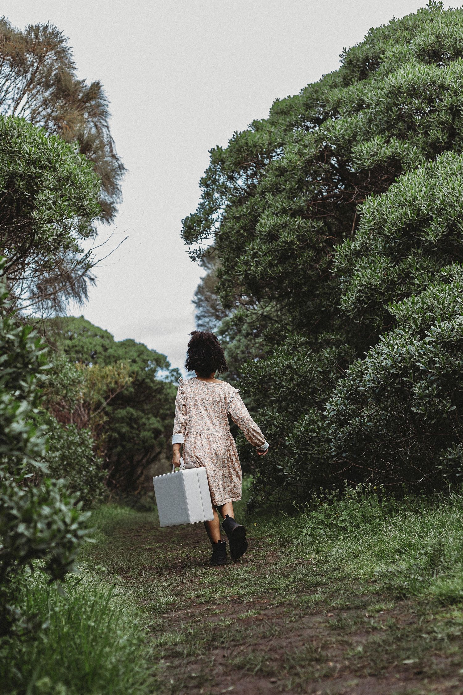 Woman in floral dress carrying a suitcase walking down a lush green forest path.