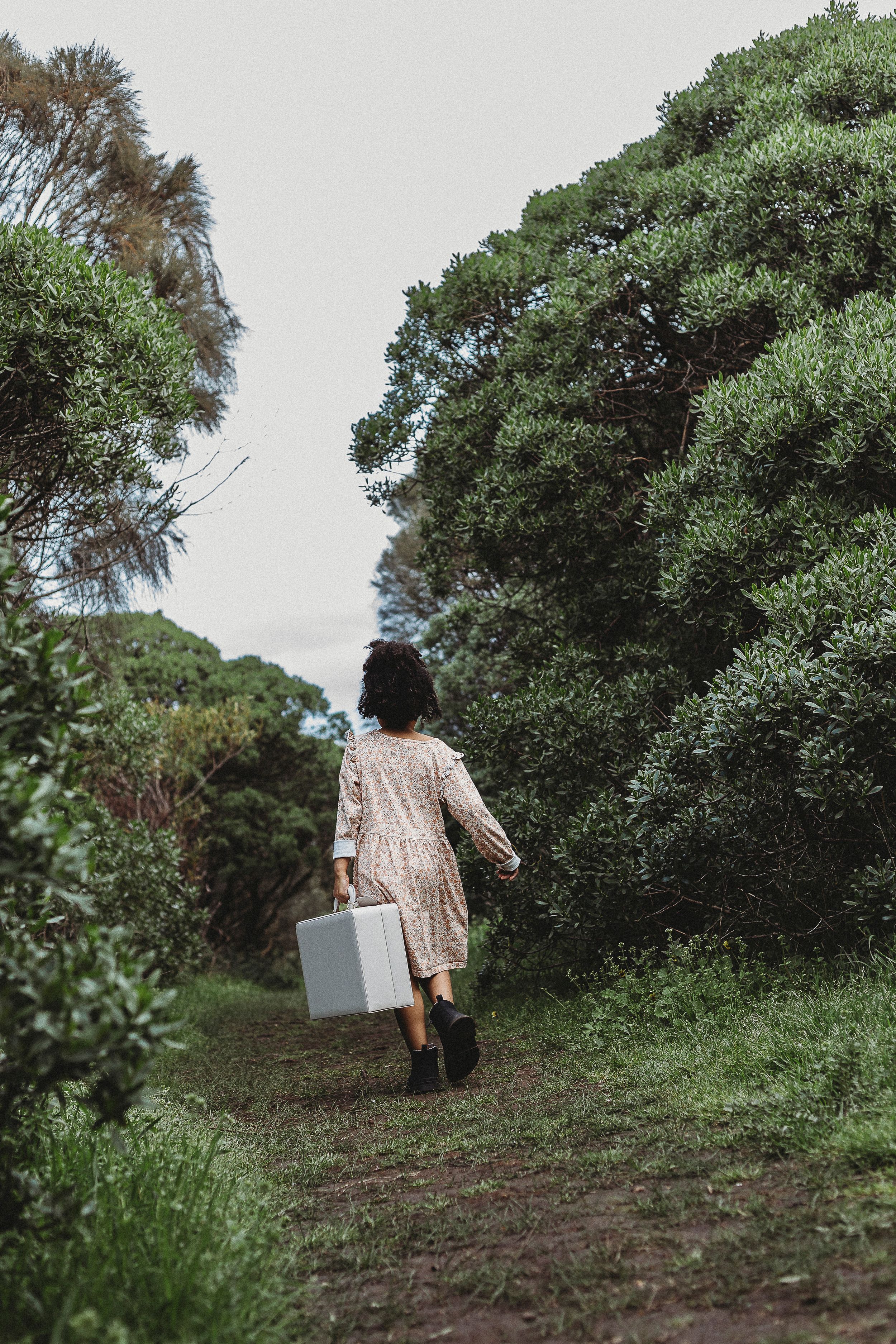 Woman in floral dress carrying a suitcase walking down a lush green forest path.