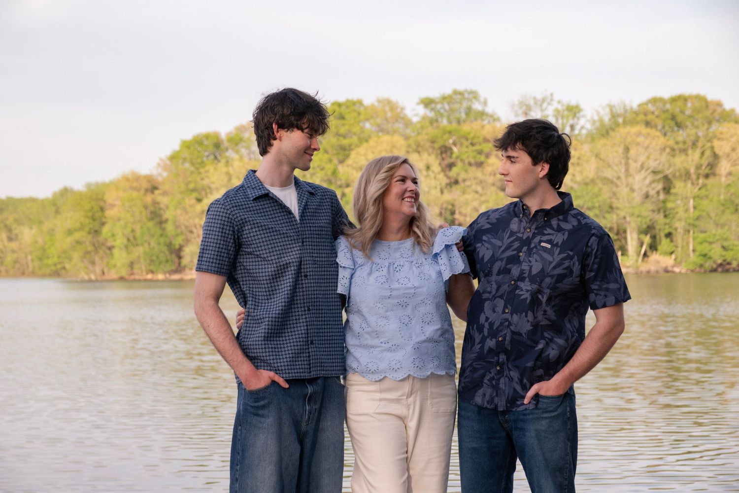 A woman stands smiling between two young men near a lake with lush green trees in the background.