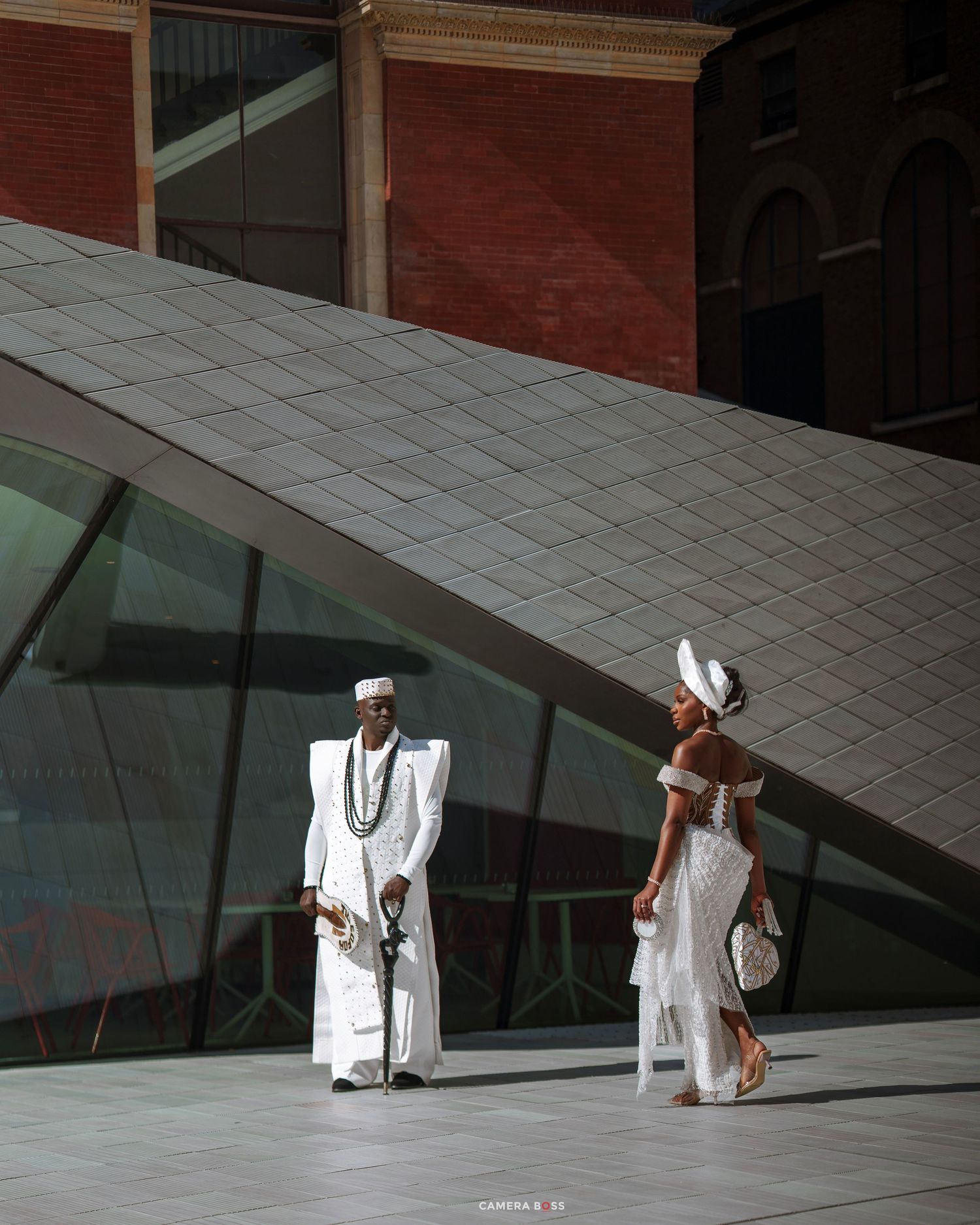 A couple in elegant white traditional African attire poses outside a modern glass building with geometric architecture.