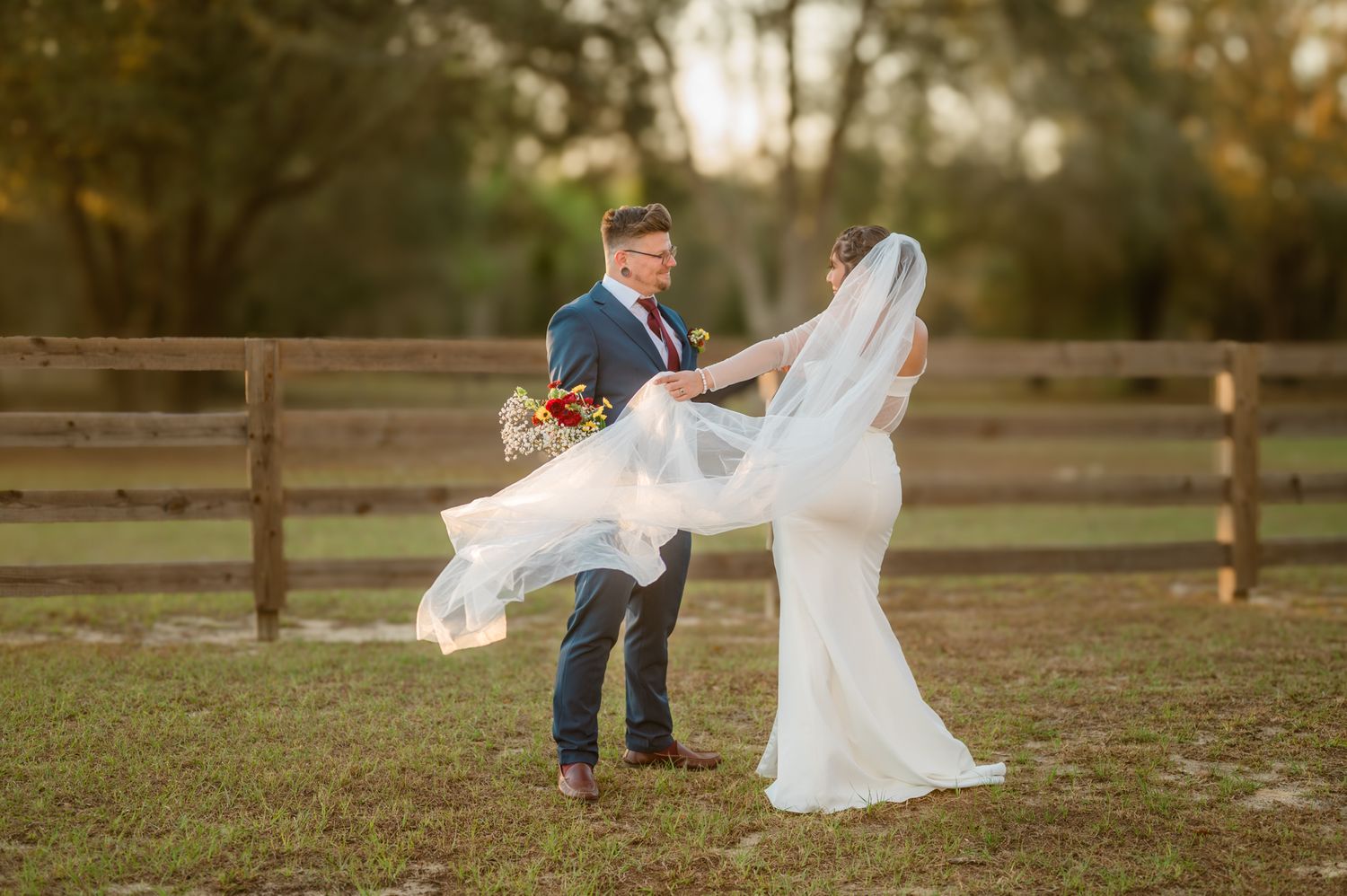 A couple in wedding attire dances outdoors with flowing veil movement against a rustic fence backdrop at sunset.