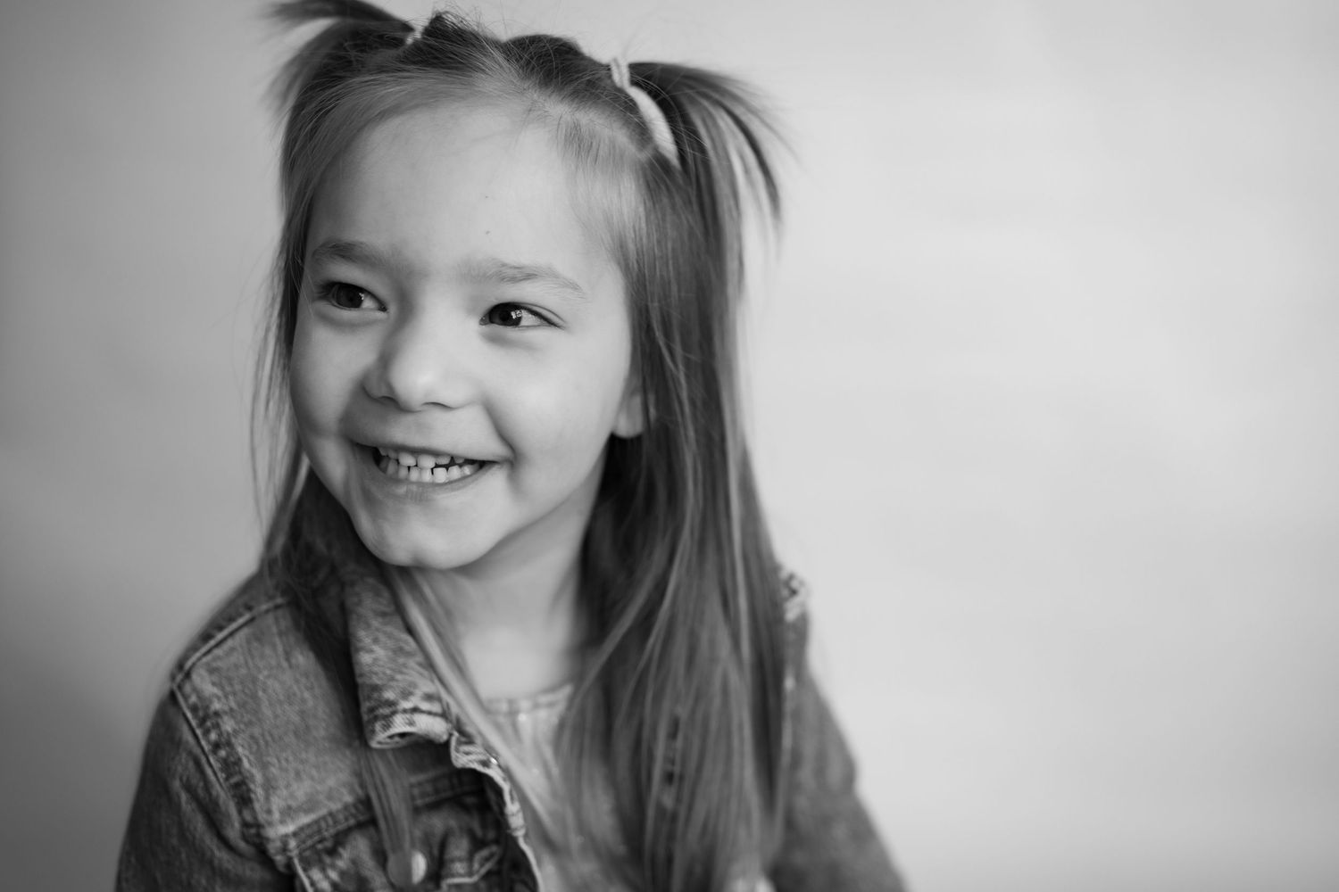 A black and white portrait of a smiling girl with pigtails wearing a denim jacket.