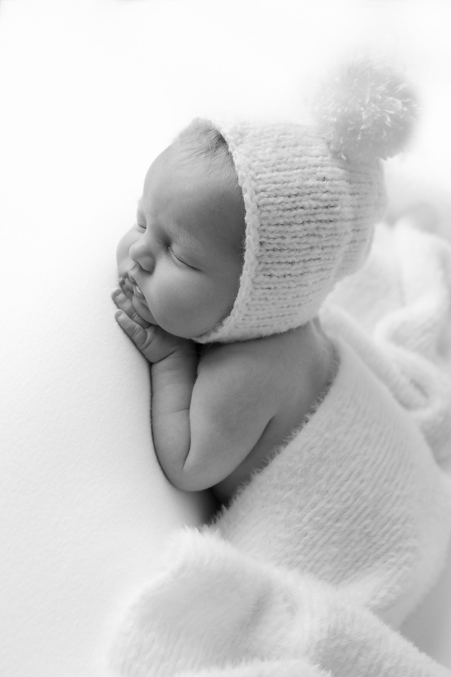 A series of black and white photos showing a sleeping newborn baby wearing a soft white knit hat against white fabric.
