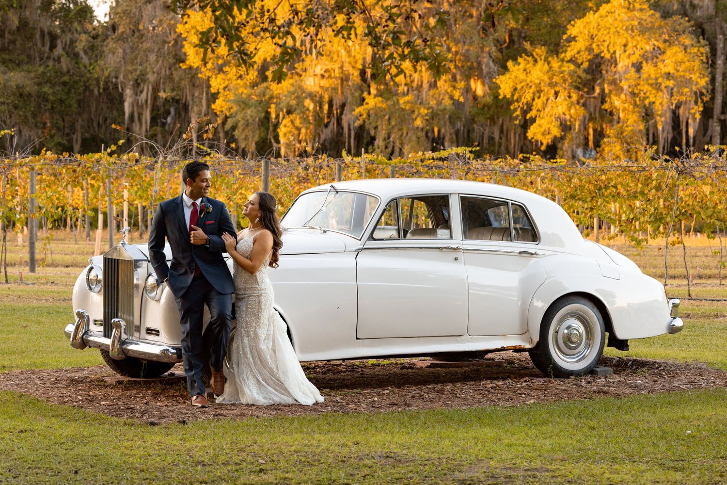 Elegant white vintage car parked in autumn setting with golden foliage and wedding couple in background.