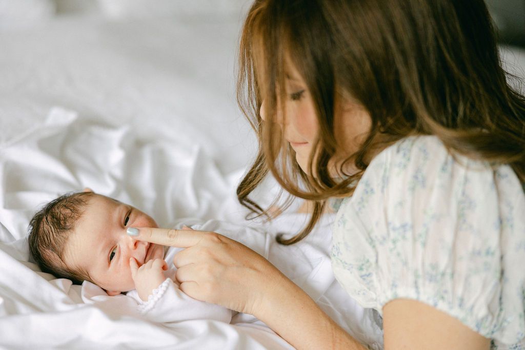 A parent and baby share a tender moment together while laying on white bedding. In-home newborn photography by Ashley Nielsen Studio.
