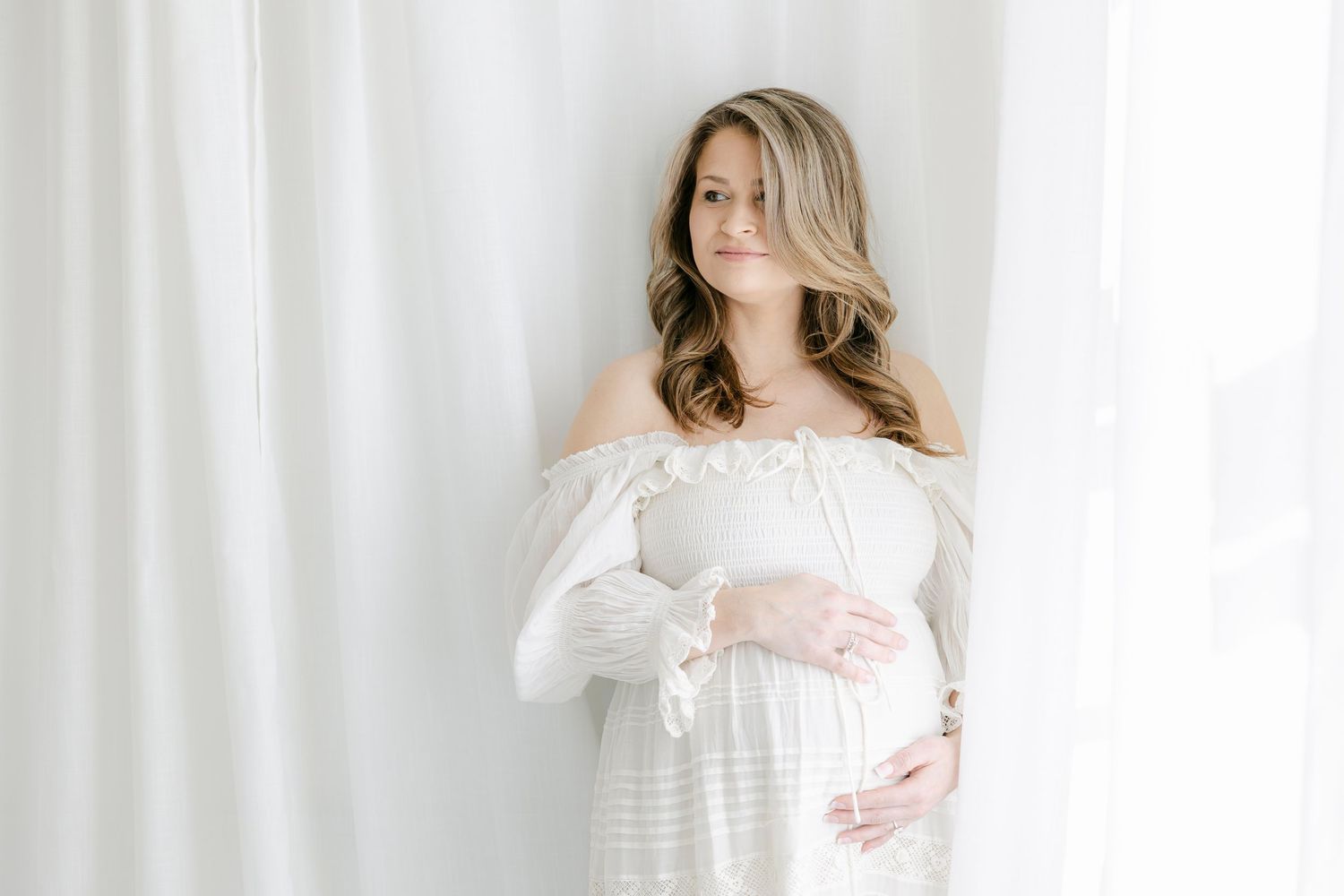 Pregnant woman in white off-shoulder dress cradling baby bump near sheer curtains in bright studio.