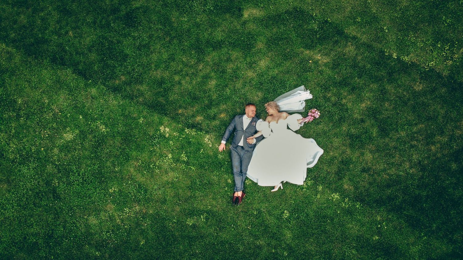 Aerial view of bride and groom lying on lush green grass, wedding dress flowing, captured from above.