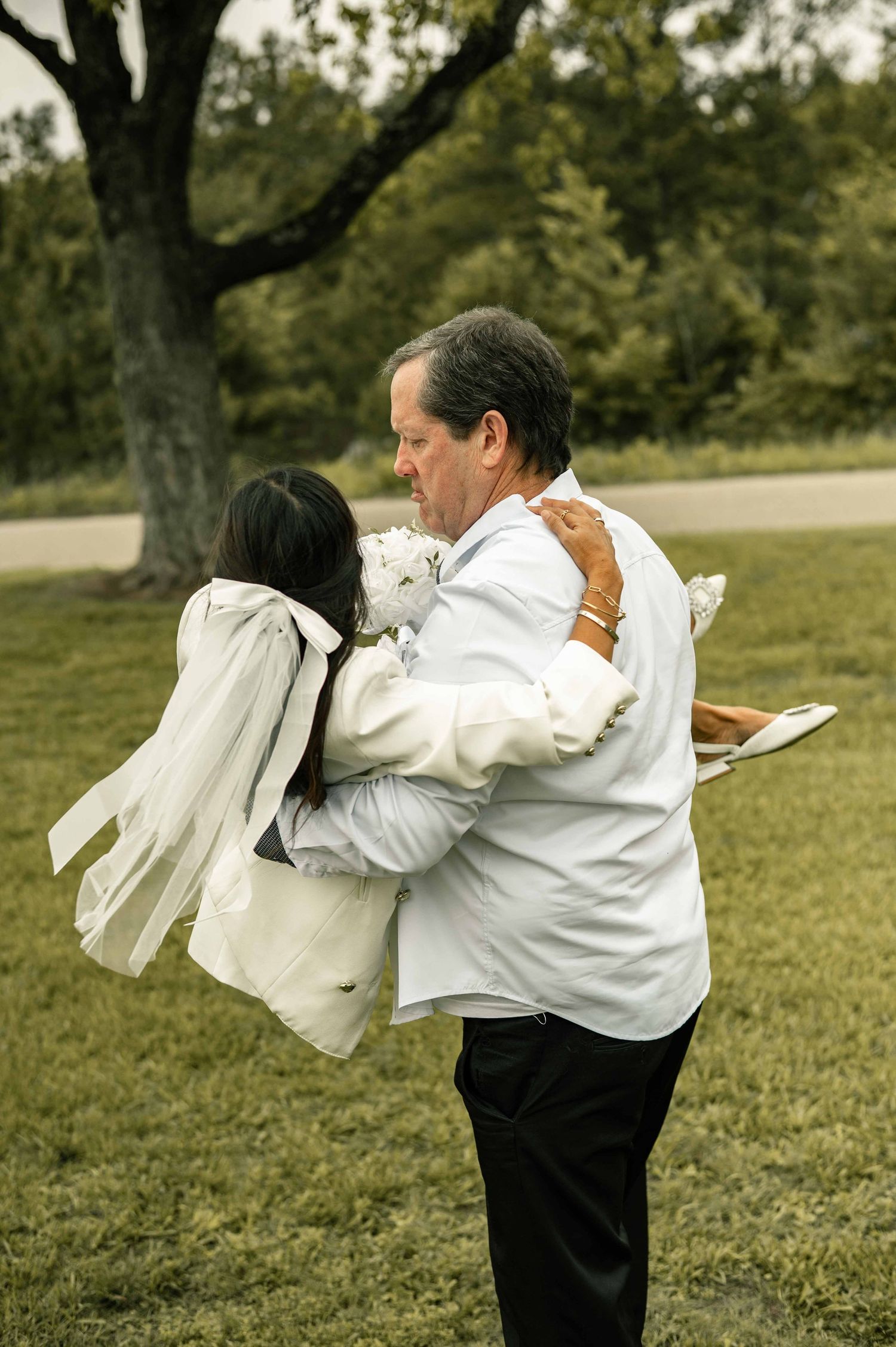 A bride and older man share an emotional embrace outdoors, surrounded by lush green trees at a wedding ceremony.