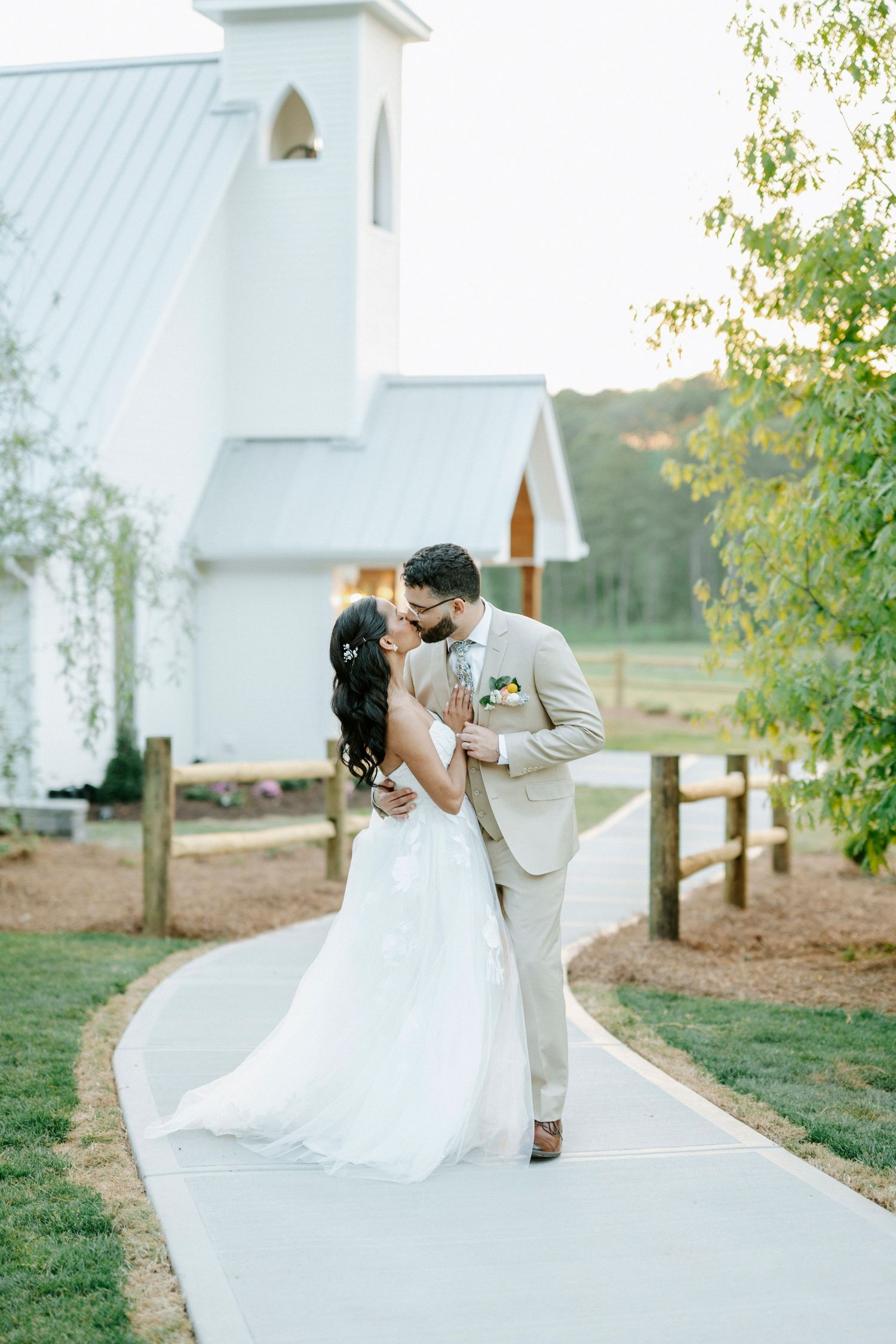 Bride and groom share a romantic kiss on a pathway outside a white chapel surrounded by lush greenery.