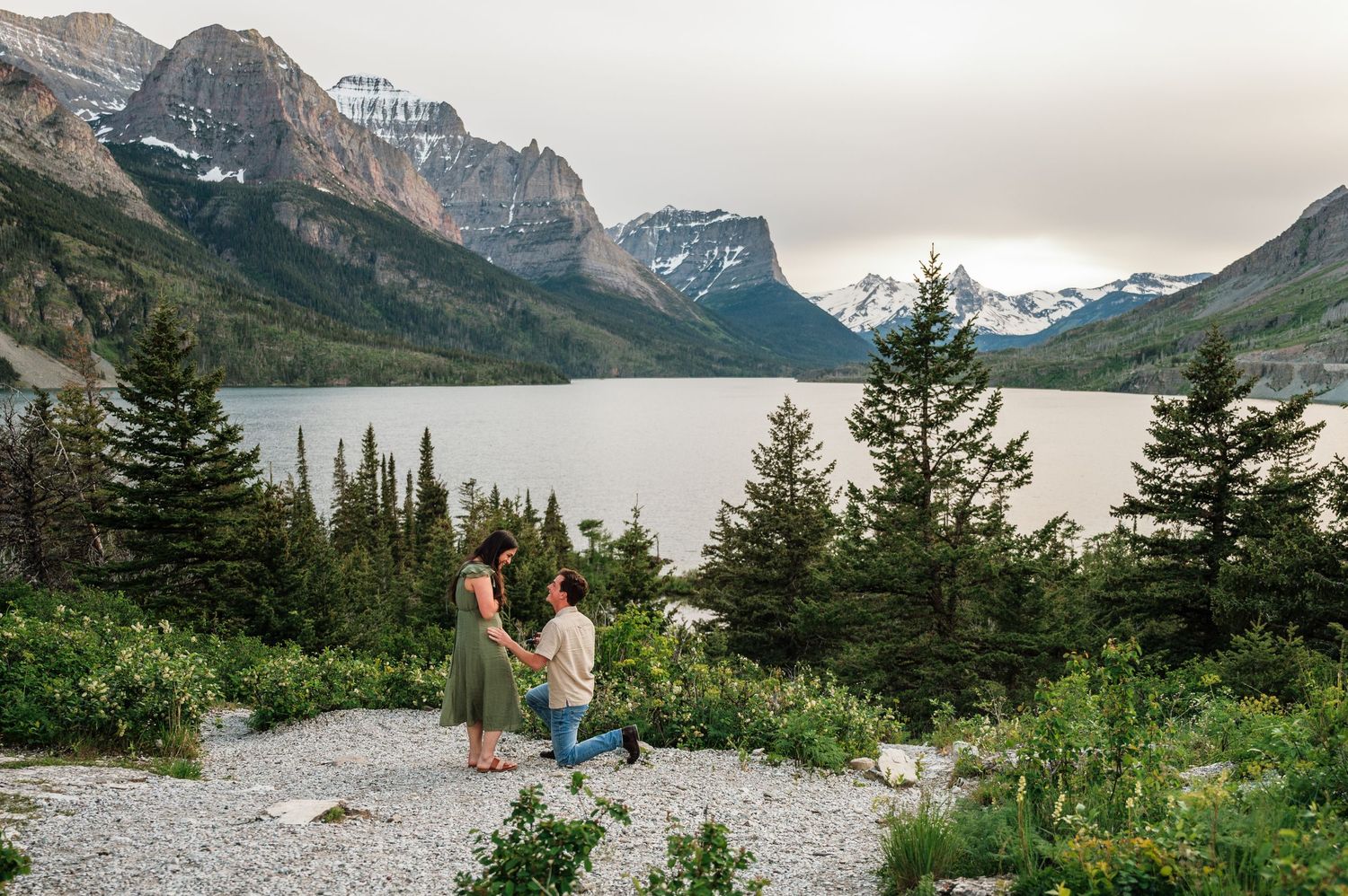 Couple sharing a marriage proposal moment by a mountain lake with dramatic peaks and evergreen trees in the background.