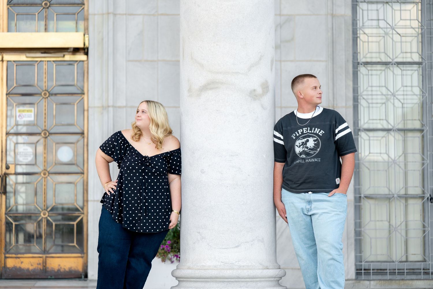 Couple posing on either side of a marble column outside a classical building, woman in polka dot top and man in graphic tee.