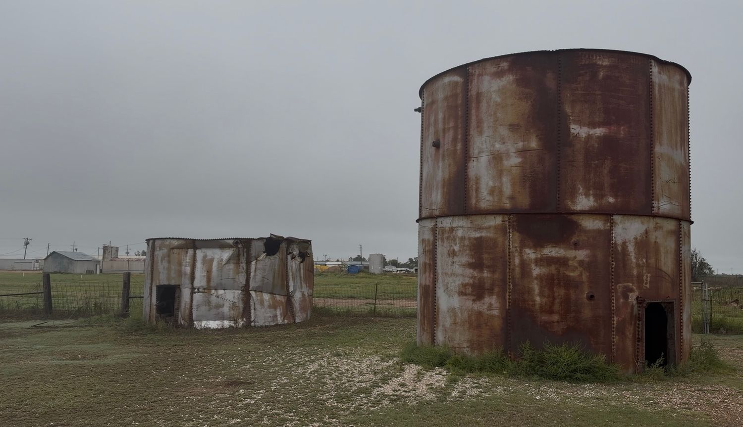 Rusty abandoned metal storage tanks on a flat grassy field under an overcast sky in a remote rural area.