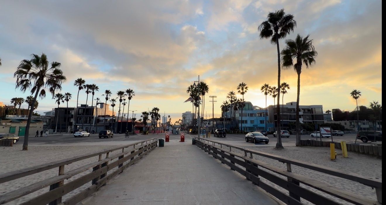 Sunset view down a wooden boardwalk lined with tall palm trees at Venice Beach, California, with colorful clouds in the sky.