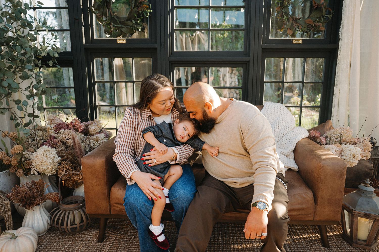 A loving family of three sits on a brown leather sofa surrounded by autumn floral decor, parents kissing their baby.