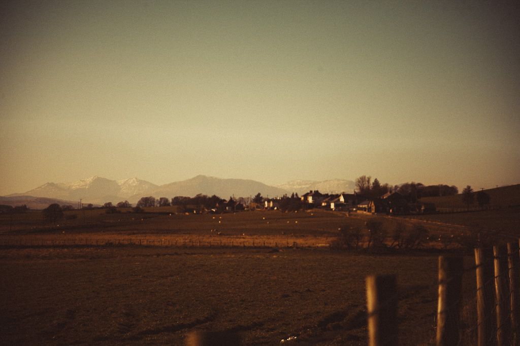 Rural farmhouses at dusk with snow-capped mountains of snowdina in background, golden fields and wooden fence posts in foreground.