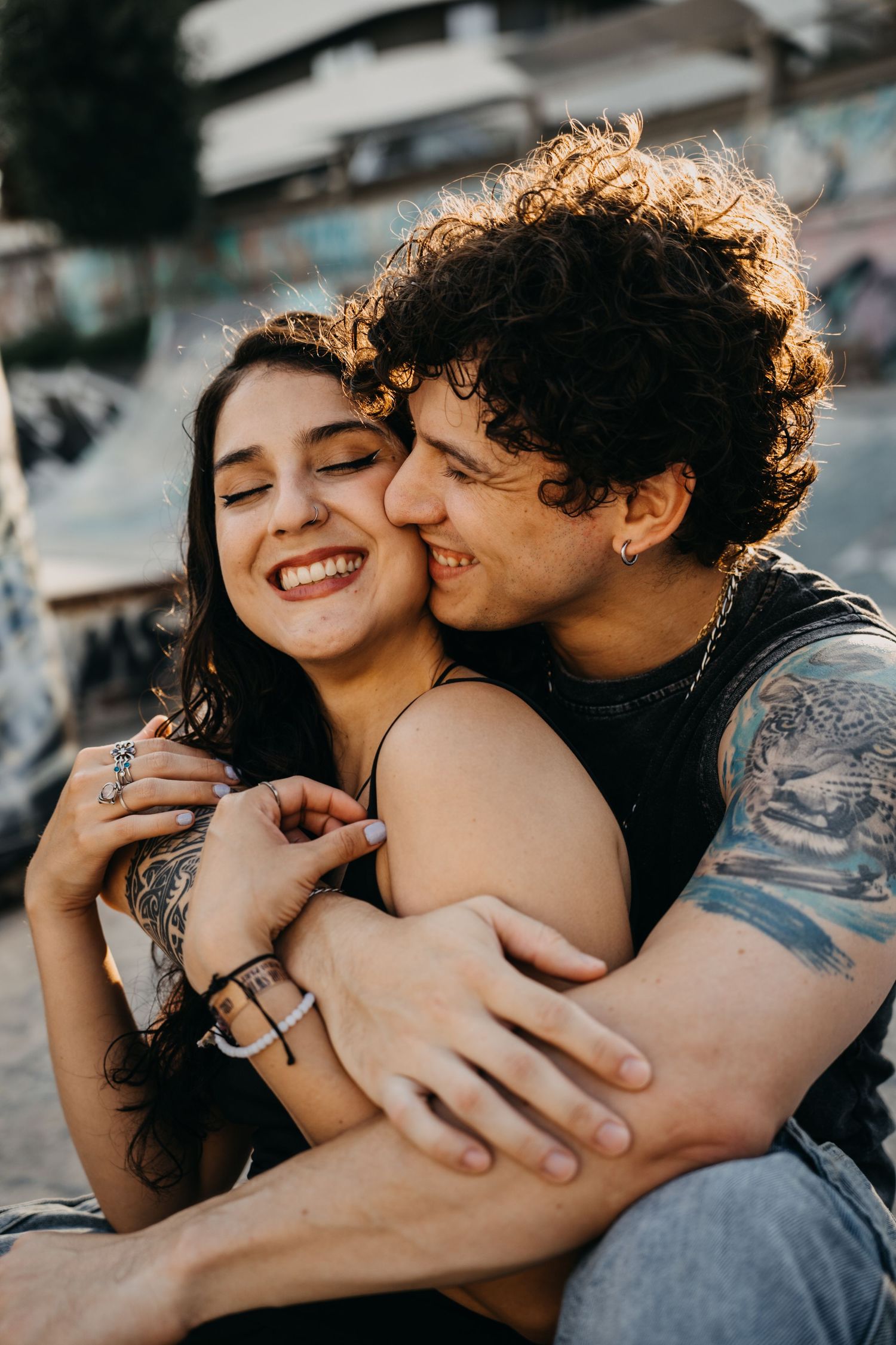 Young tattooed couple embracing and smiling outdoors, man kissing woman on cheek in warm golden sunlight.