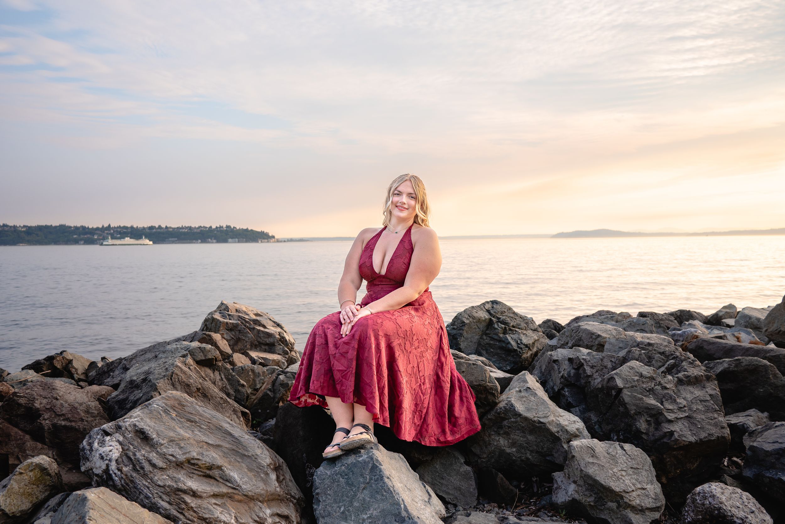 High school senior in a burgundy dress posing on shoreline rocks at Olympic Sculpture Park in Seattle