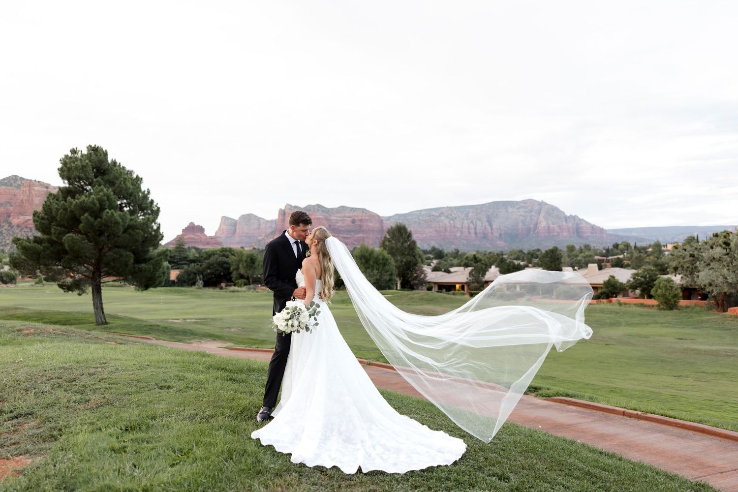 Bride and groom kissing on a green lawn with stunning red rock Sedona Arizona mountains in the background, veil flowing in wind.