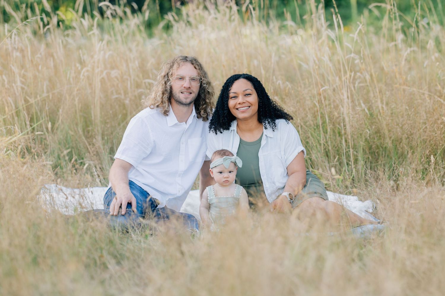 A happy interracial family with a baby sits in a golden field of tall grass for an outdoor portrait.