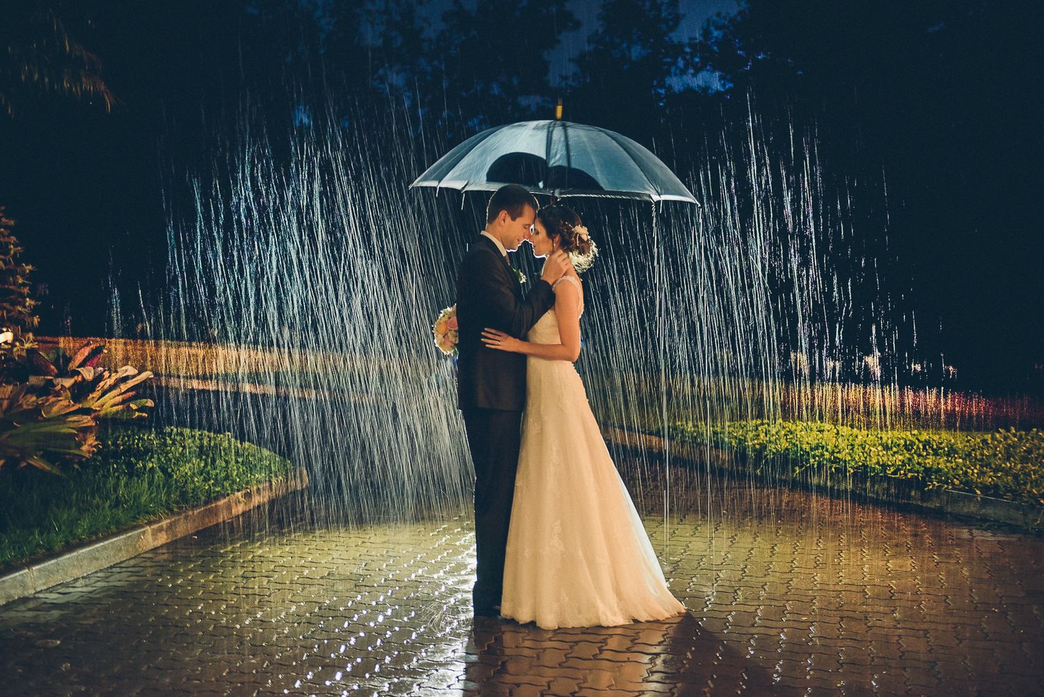 Couple embraces under umbrella in rain with dramatic lighting creating magical wedding moment.