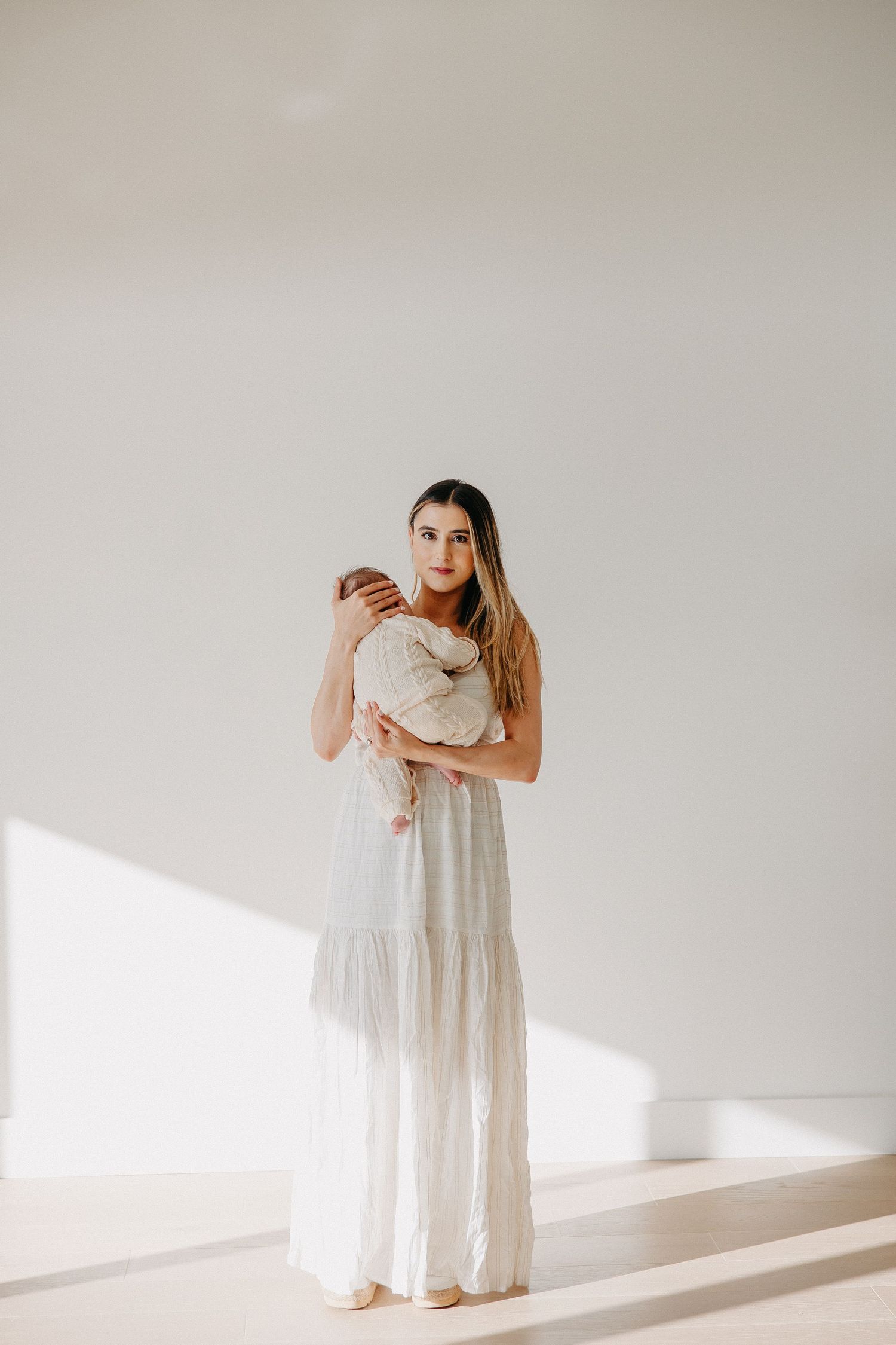 A person in a flowing white maxi dress stands against a minimalist white wall in natural sunlight.