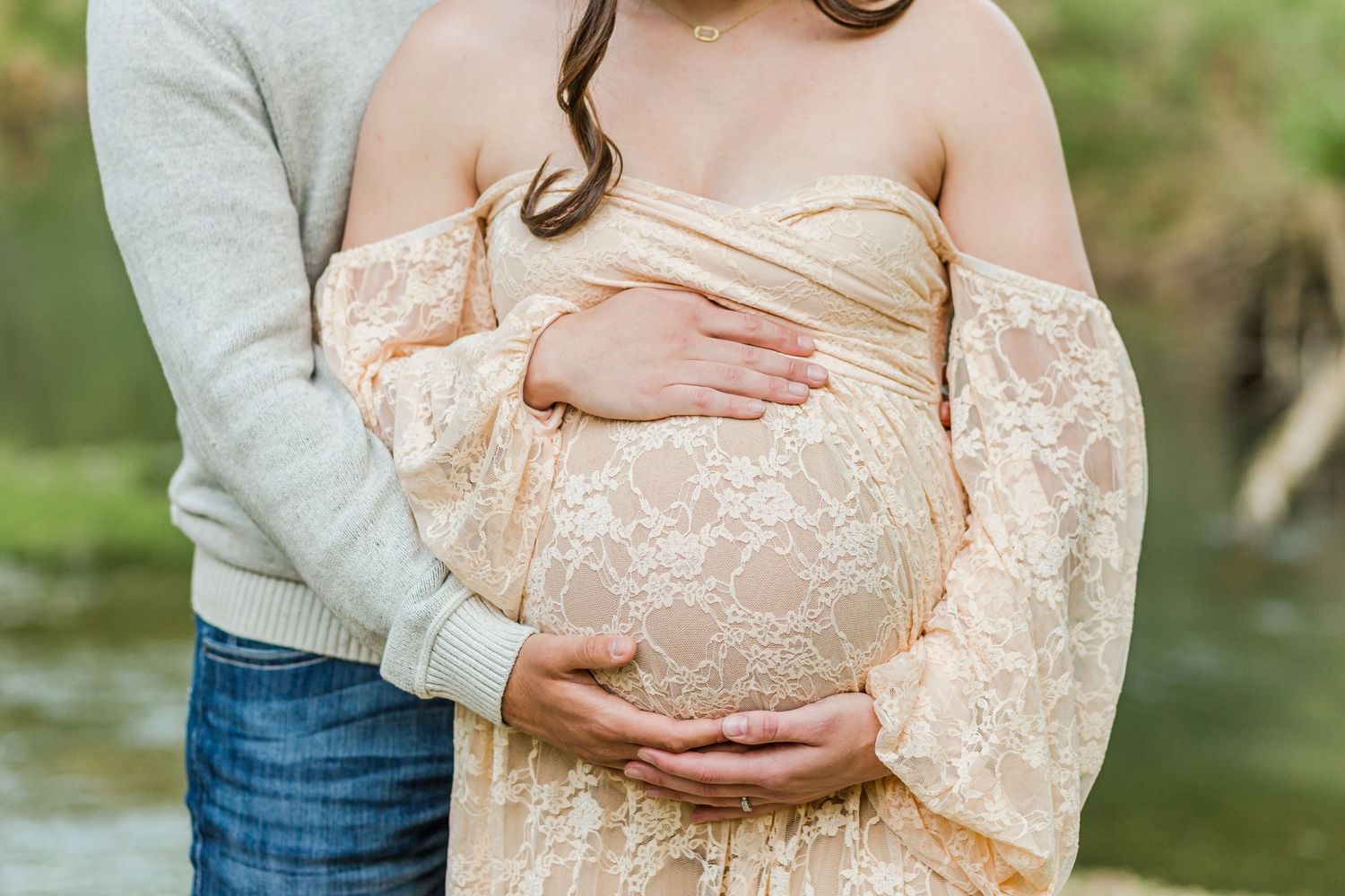 Couple shares intimate moment highlighting a baby bump in a lace maternity dress against nature backdrop.