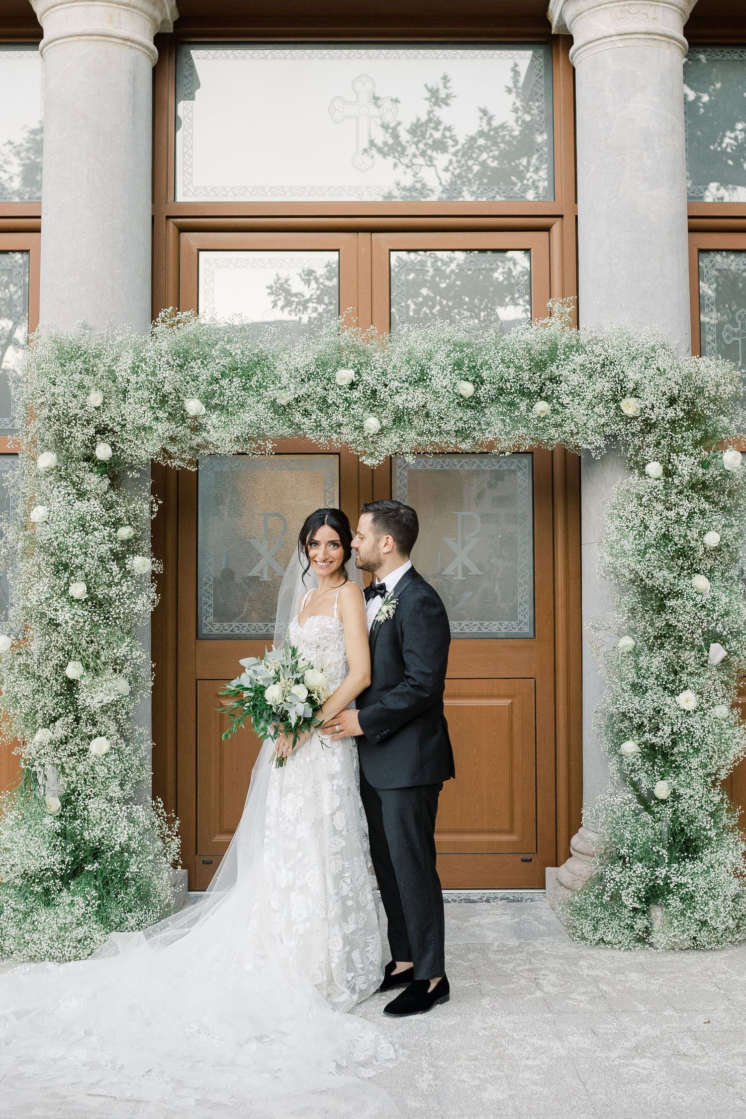Wedding couple stands under floral arch with baby's breath against wooden doors.
